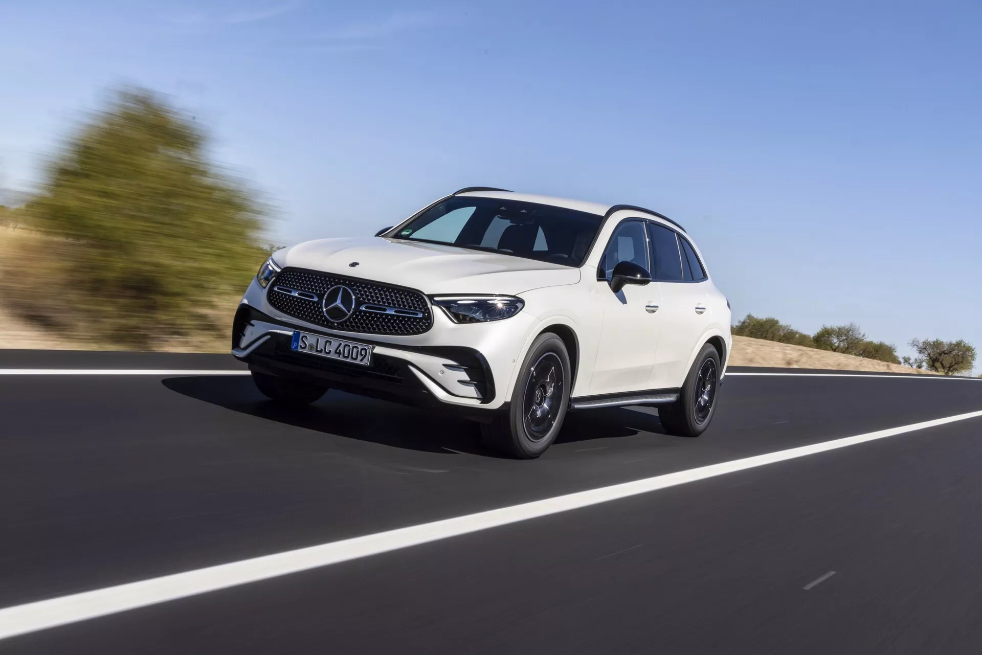 Mercedes GLC 2022 (254) vit fyrdörrars crossover SUV i rörelse på öppen väg, fångad från en låg vinkel som betonar dess höjd och hållning. Fordonet rör sig diagonalt från vänster till höger, med fokus på främre grillen, strålkastarna, LED-varselljus och nedre främre stötfångaren. Sidprofilen avslöjar den skulpterade taklinjen, designlinjerna, de utställda skärmkanterna och lättmetallfälgarna, som kombinerar lyx och sportighet. Den suddiga bakgrunden antyder hastighet, under en klar dagsljushimmel.