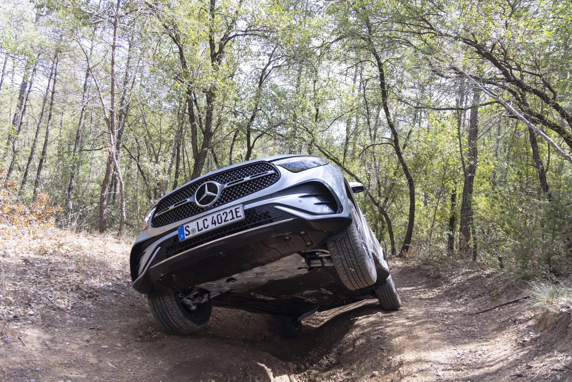 2022 Mercedes-Benz GLC 254 fångad i en offroadmiljö i en skog som visar dess förmågor i tuff terräng. Den låga, något framåtvinklade bilden belyser fordonets frontgrill, skyddsplåt och fjädringsartikulation med vänsterhjulen upphöjda över en kulle och högerhjulen i en dike. Detta betonar bilens markfrigång och offroad-skyddsegenskaper. EQ-märket antyder el- eller hybridteknik. Naturligt dagsljus, ingen artificiell belysning.