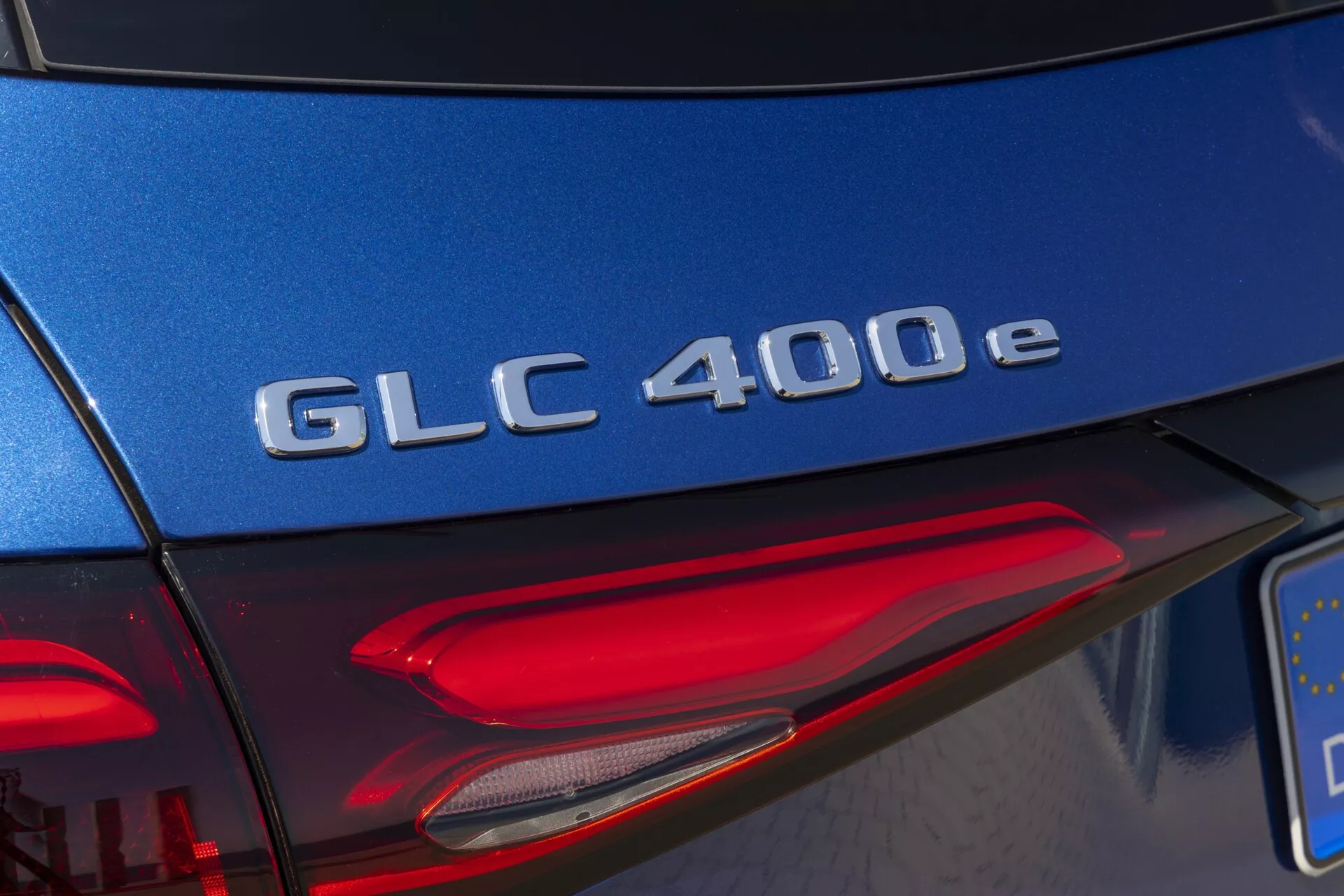 Detalj av bakre sektionen på en Mercedes-Benz GLC 400e 2022, med en närbild av modellbeteckningen i krom på en blå baklucka, med en diagonal vinkel som fångar en del av den vänstra bakljusenheten med LED-element i dagsljus.