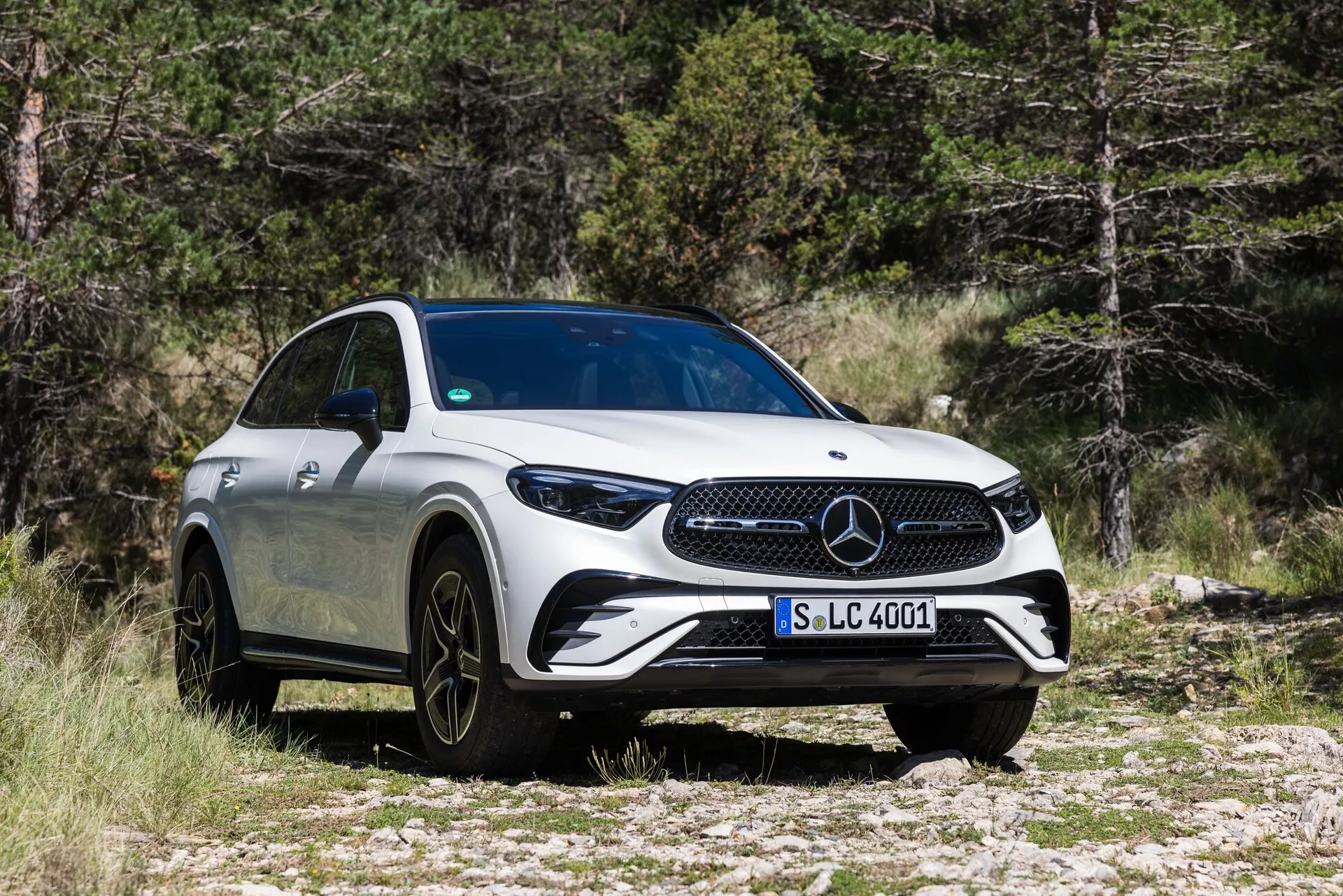Trekvarts främre vy av en Mercedes-Benz GLC 2022, som framhäver dess framträdande grill, eleganta LED-strålkastare, stora multi-ekrade svarta aluminiumfälgar och aerodynamiska vita kaross. Fordonet är parkerat på stenig terräng med träd i bakgrunden, vilket betonar dess SUV-förmågor och lyxiga utseende.
