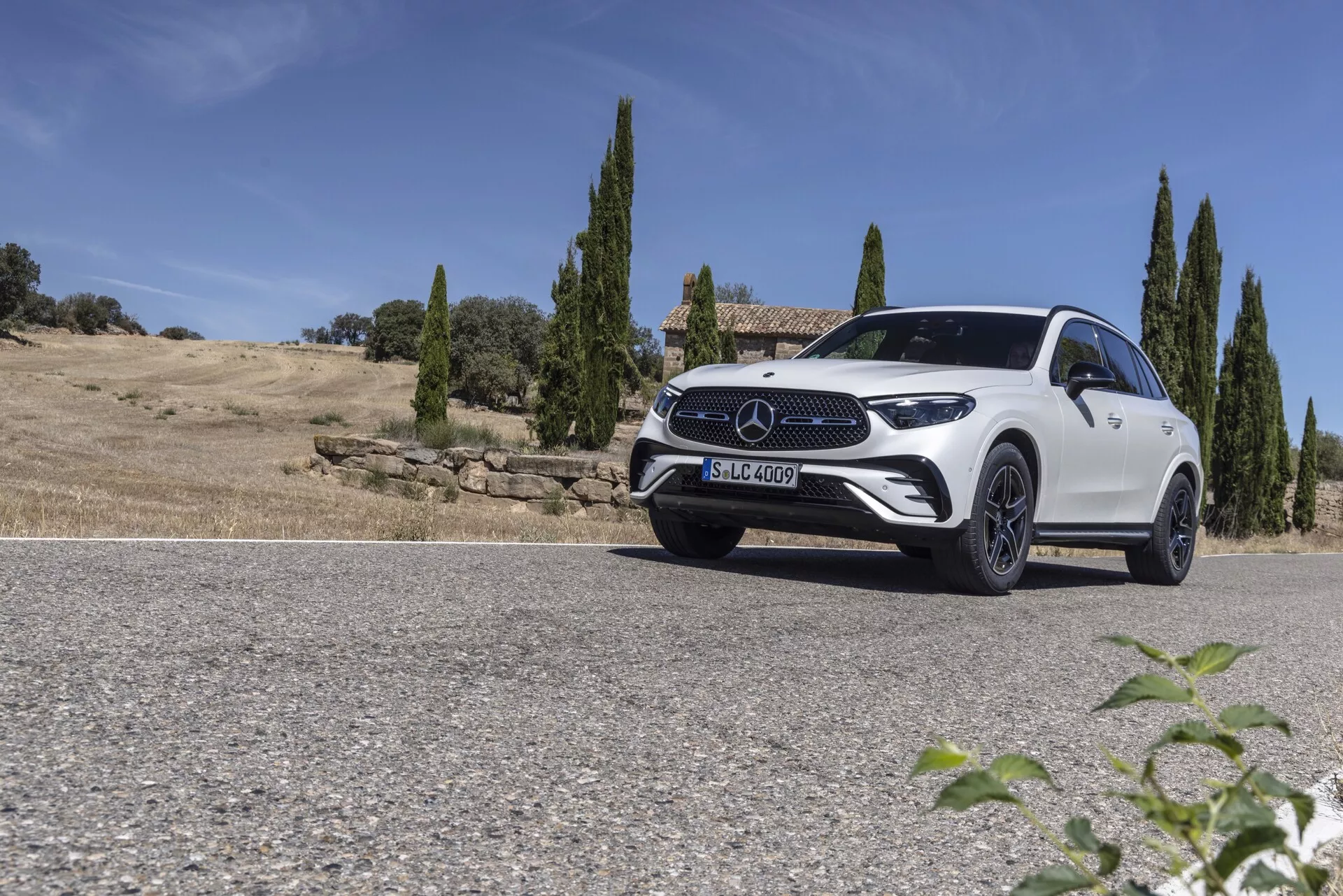 Låg vinkelbild av en vit Mercedes GLC 2022 SUV parkerad på en väg mot en naturlandskap med klar himmel och cypressträd i bakgrunden. Fordonets främre kvarts fångas från förarsidan och visar de eleganta LED-strålkastarna, den framträdande framgrillen med stort emblem, sportiga legeringshjul och den konturerade huvan. Den något sluttande taklinjen och den väl definierade skuldlinjen framhäver designen som kombinerar aerodynamik och robusthet. Det starka solljuset skapar skuggor och höjdpunkter som förstärker bilens lyxiga och moderna estetik.