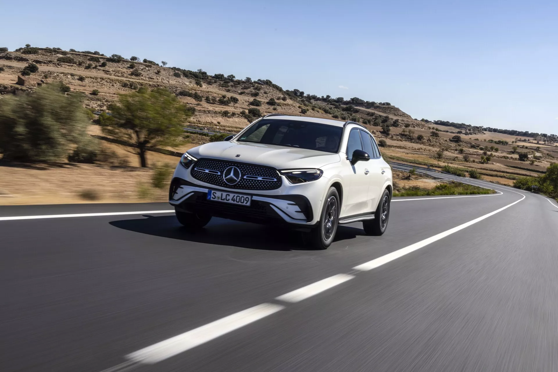Dynamisk låg vinkelbild av en vit Mercedes-Benz GLC SUV 2022 i rörelse på en kurvig väg. Trekvartsvyn framifrån fångar grillen med Mercedes-Benz-emblemet, strålkastarna och de främre stänkskärmarna. Den slanka aerodynamiska designen och lättmetallfälgarna framhäver fordonets hastighet och lyx. Sett i en ökenliknande miljö med gles vegetation, tyder bilens integrerade främre sensorer och sidospeglar på avancerade förarassistans- och säkerhetssystem.