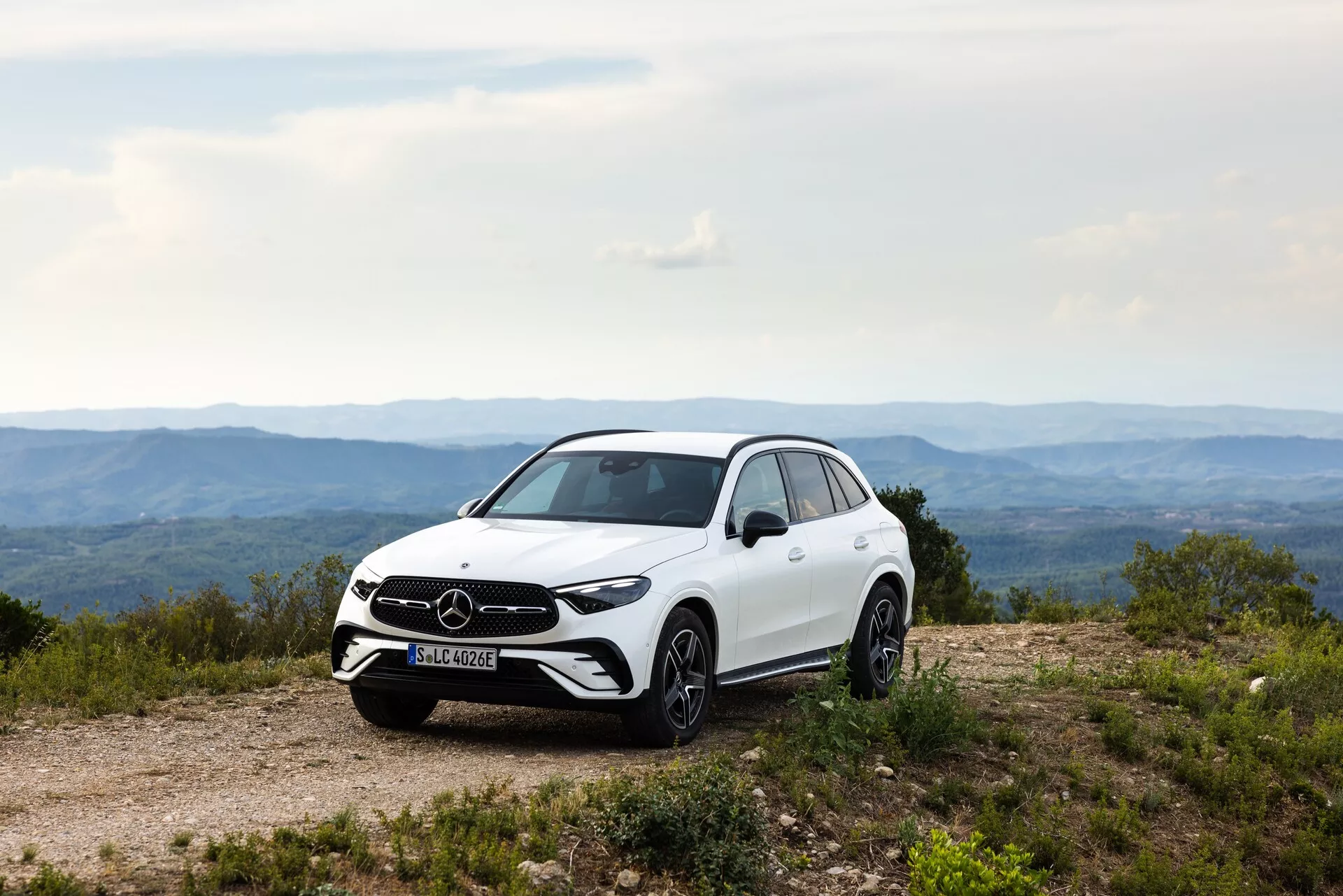 Sidoprofil av vit 2022 Mercedes GLC på en ojämn grusväg, som framhäver SUV:n:s hållning, markfrigång och designdetaljer. Synliga funktioner inkluderar frontgrill och varumärkesemblem, LED-strålkastare, aluminiumfälgar, sidobackspeglar med integrerade blinkers, aerodynamiska karosslinjer, blank färgfinish och däckens slitbanemönster. Det naturliga dagsljuset framhäver bilens estetiska och funktionella egenskaper.