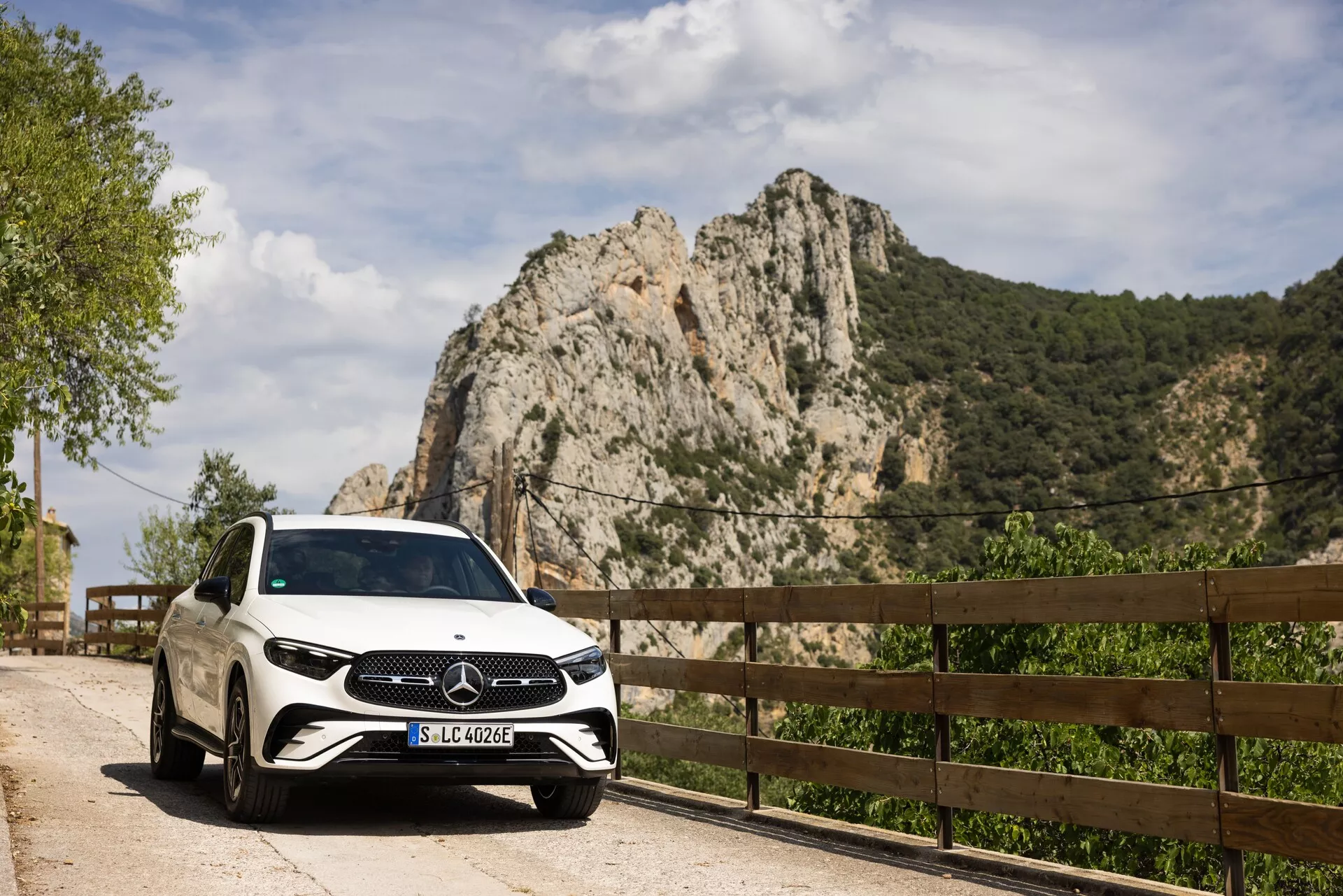 Vit Mercedes-Benz GLC 2022 parkerad vid en naturskön väg med en framifrån-höger vy som visar grill, strålkastare och delar av höger sida inklusive främre passagerardörr och hjul. Registreringsskylt 'SLC 400 e' som indikerar möjlig plug-in hybrid eller elfordon. Fotograferad under dagsljus, bilens vita lack och blanka yta reflekterar naturligt ljus och framhäver det stora Mercedes-Benz-emblemet på grillen. Klippig berg och trästaket i bakgrunden antyder en lantlig eller bergsväg, vilket tyder på en lyxig reträtt eller utomhusutforskning.
