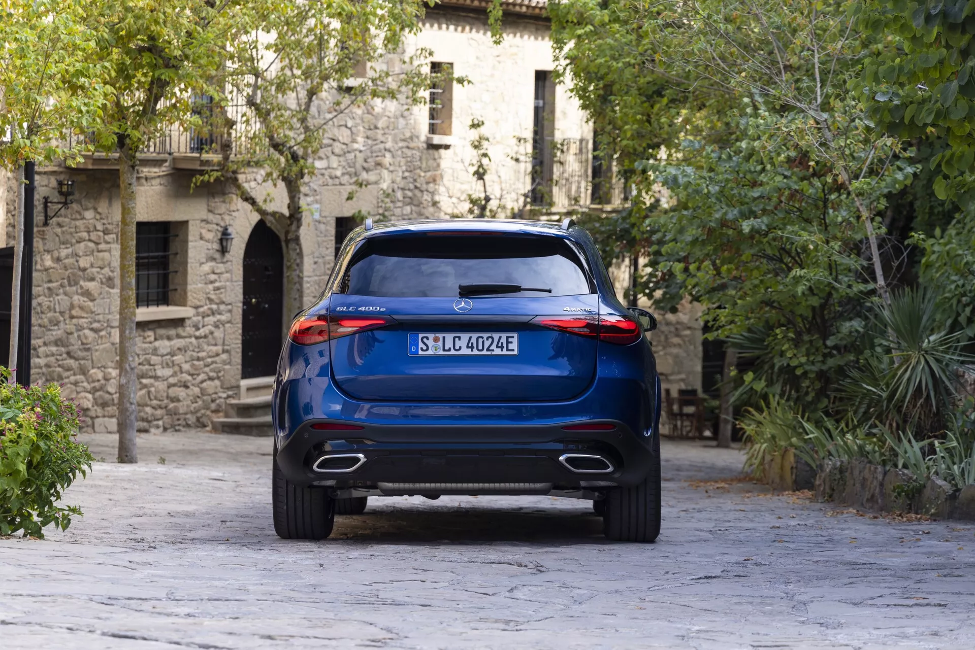 Bakre vinkelvy av en blå Mercedes GLC 2022 SUV från en något låg perspektiv, som visar dess höga markfrigång, dubbla avgasutlopp och eleganta bakljuskluster. Fordonet, placerat centralt, står mot en bakgrund av europeisk arkitektur och kullerstensväg, vilket framhäver dess modernitet och möjliga elektriska drivlina.