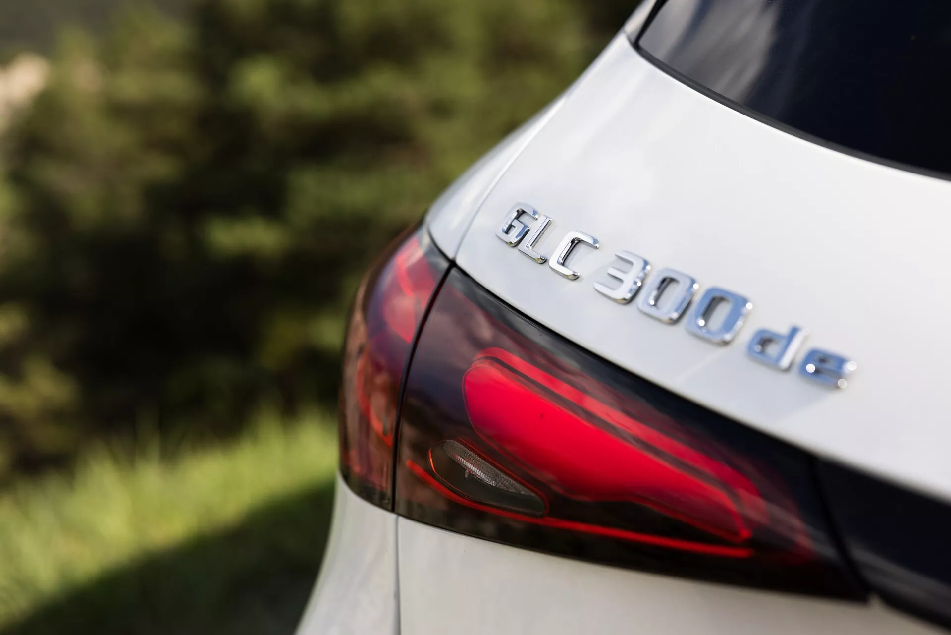 Närbild av den bakre delen av 2022 års Mercedes GLC 300de, med fokus på modellbeteckningen och en del av LED-baklyktan. Bilden, tagen från en något upphöjd sidvinkel, framhäver GLC 300de-emblemet och den släta konturen av den moderna baklyktdesignen. Den metalliska lacken reflekterar ljuset subtilt, vilket tyder på en välunderhållen yta, även om den exakta färgen verkar vara ljus, möjligen vit eller silver. Det grunda skärpedjupet håller emblemet i skarp fokus samtidigt som bakgrunden och de perifera områdena suddas ut, vilket betonar de tekniska och designmässiga detaljerna hos detta diesel-elektriska hybridfordon.