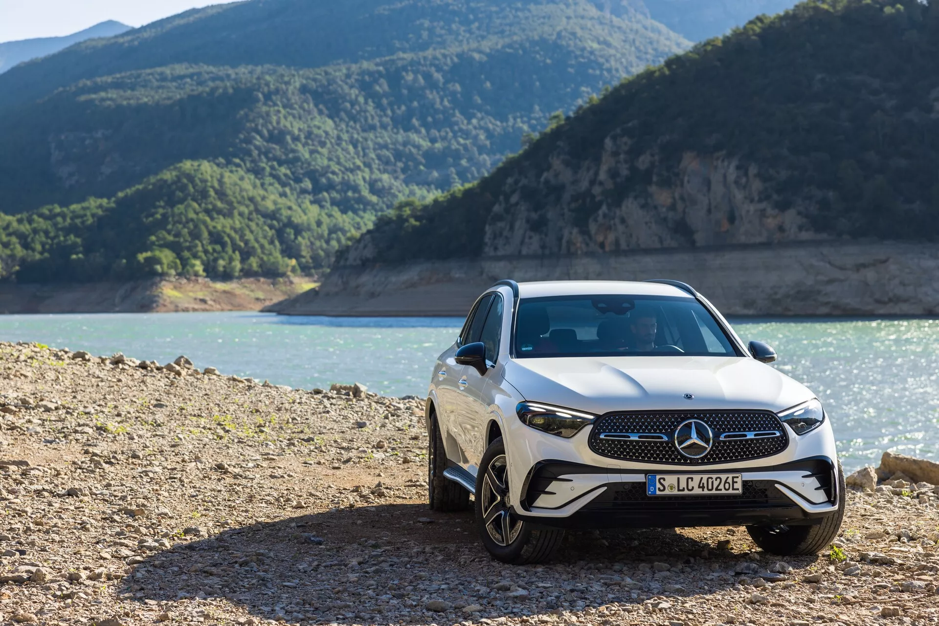 Främre tredjedels vy av en lyxbil Mercedes GLC 254 från 2022, som visar dess främre grill med framträdande emblem, eleganta LED-strålkastare och lättmetallfälgar, parkerad i en robust utomhusmiljö nära en strandlinje med berg i bakgrunden.