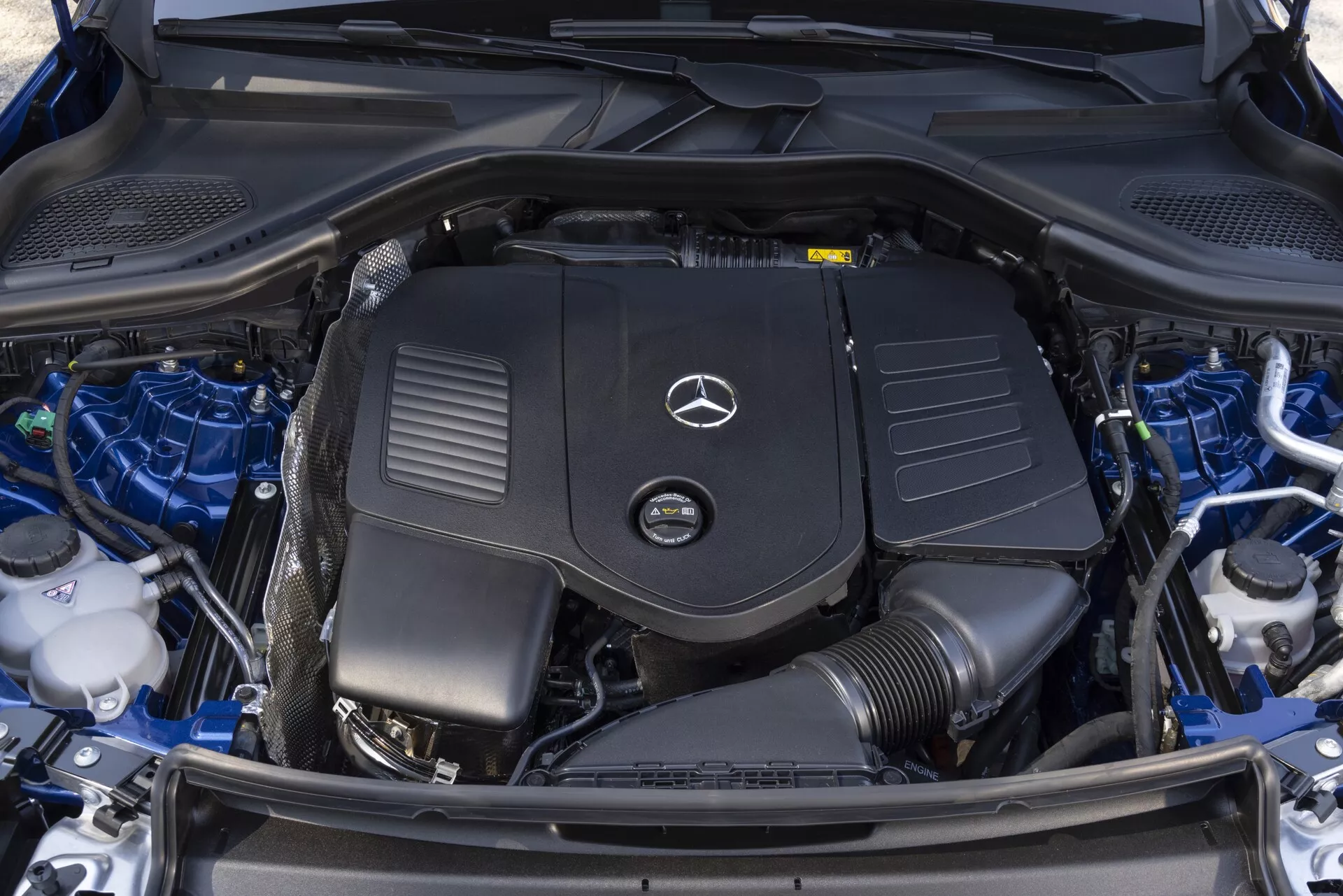 Närbild från ovan av motorrummet i en 2022 Mercedes GLC, som är rent och välskött, med motorhuven öppen och motorlocket med Mercedes-Benz-logotypen tydligt synligt, batteri, vätskebehållare, kylsystemkomponenter och prydligt organiserade kablage.