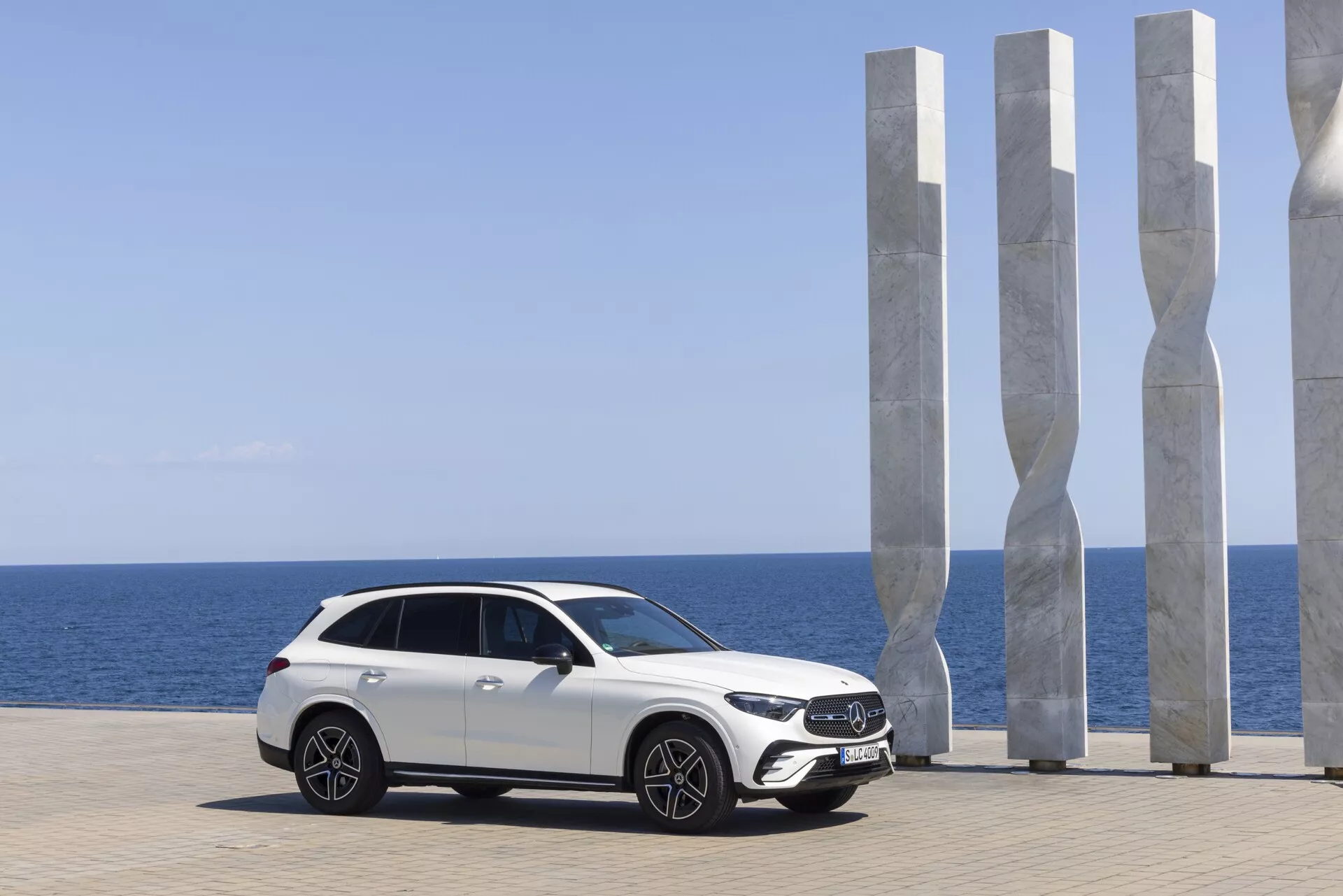 Modern vit lyxig Mercedes GLC 2022 parkerad med naturskön kustutsikt; sidoprofil som visar elegant design med hög markfrigång, stora lättmetallfälgar, avancerade LED-strålkastare, aerodynamiska speglar med integrerade blinkers, sluttande kupélinje, panoramasoltak och tonade rutor. Natursköna bakgrunden med moderna vita skulpturer, klarblå himmel och lugnt hav förstärker den premiumautomotiva tilldragelsen.
