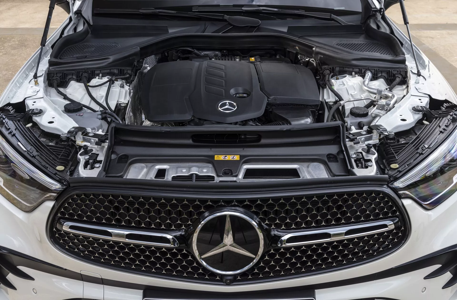 Närbild av fronten på Mercedes-Benz GLC 2022 (254) med huven öppen som visar motorutrymmet från ögonhöjdsperspektiv, centrerad på märkets emblem och grillen, vilket framhäver den stora diamantmönstrade grillen, Mercedes-Benz stjärnemblem, motorutrymmet med logotypen och synliga komponenter som motorblock, luftintagssystem, kylvätskebehållare, fjädertorn och frontkamera som indikerar avancerade tekniska funktioner.