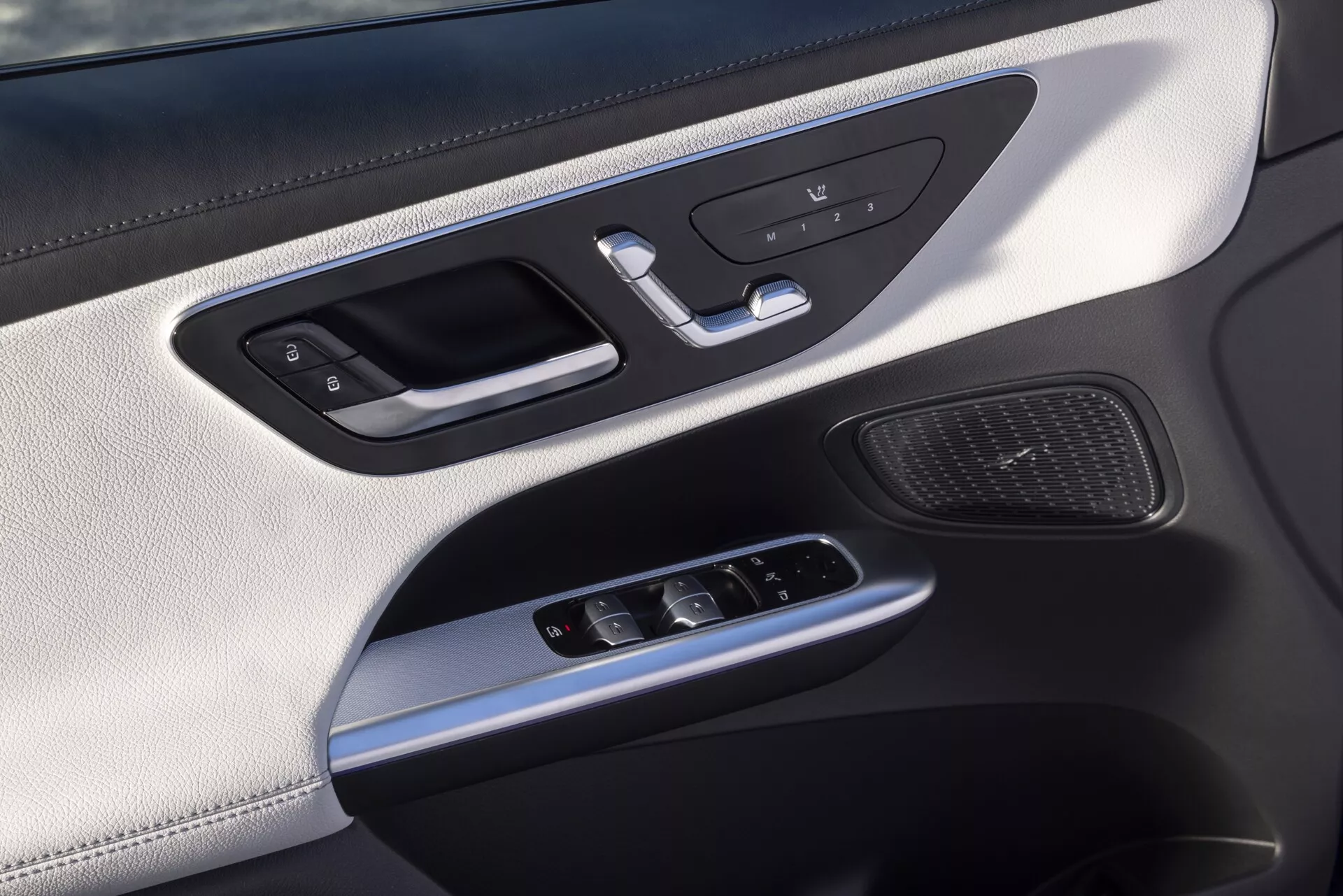 Närbild av insidan på en bildörr i en Mercedes GLC 2022 254, med fokus på förar- eller passagerarsidan, som visar en silverfärgad handtag med integrerad låskontroll, minnessätenknappar, strömbrytare för fönster, joystick för justering av sidospeglar, högtalgaller, lyxiga material inklusive en övre sektion i läderliknande material, silverfärgade detaljer och omgivande belysning.