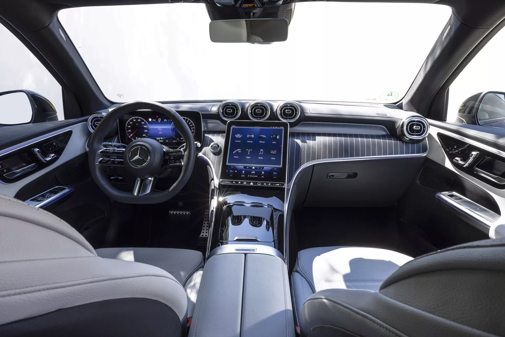Interiör av Mercedes GLC 2022, sett från centrala baksätet. Den lyxiga kabinen har en tvåtonig färgschema med krämfärgade läderklädslar och mörka accenter. Instrumentpanelen har en elegant träinredning med ett stort vertikalt pekskärms-infotainmentsystem, ventilationsöppningar och kontrollknappar. Det multifunktionella läderklädda ratten med metalldetaljer och den digitala instrumentpanelen är också synliga.