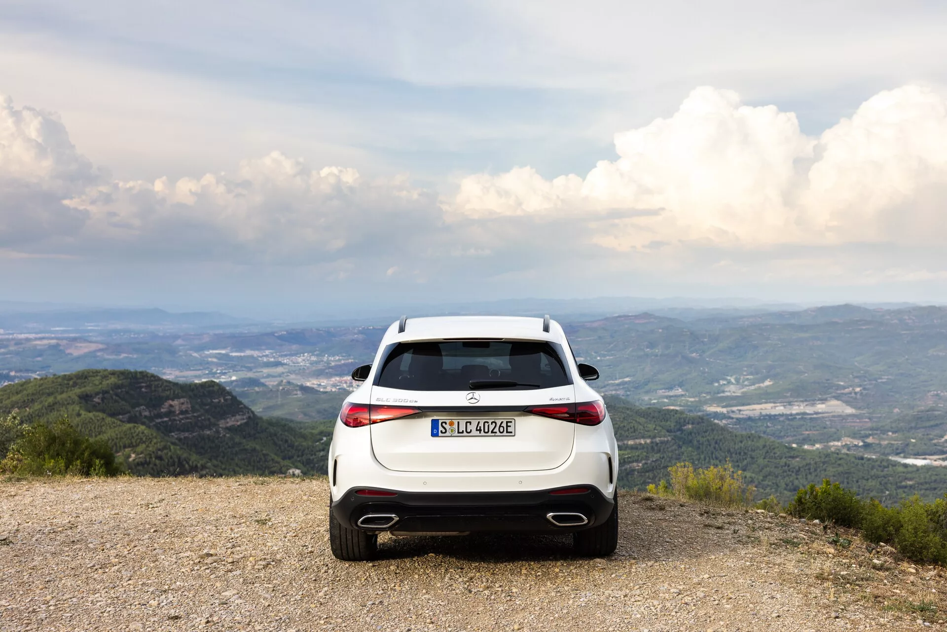 Bakifrånvy av en vit Mercedes-Benz GLC 2022 (EQ) hybrid/elektrisk SUV parkerad på en grusad yta med en naturskön bakgrund av berg och träd under en delvis molnig himmel. Fotot lyfter fram fordonets horisontellt orienterade LED-bakljus, dubbla avgasrör, bakre diffusor och EQ-märkning. Kompositionen antyder teman om äventyr och miljövänligt resande.