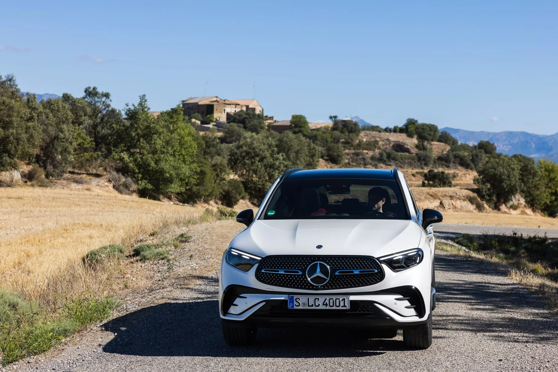 En framvinkel av en modern Mercedes-Benz GLC 2022 (254) på en smal väg, med vit bas och svarta detaljer, LED-strålkastare och en distinkt grill. Bilden tagen i ögonhöjd under dagsljus visar bilens sportiga och premium trim med ett medelhavslandskap i bakgrunden.