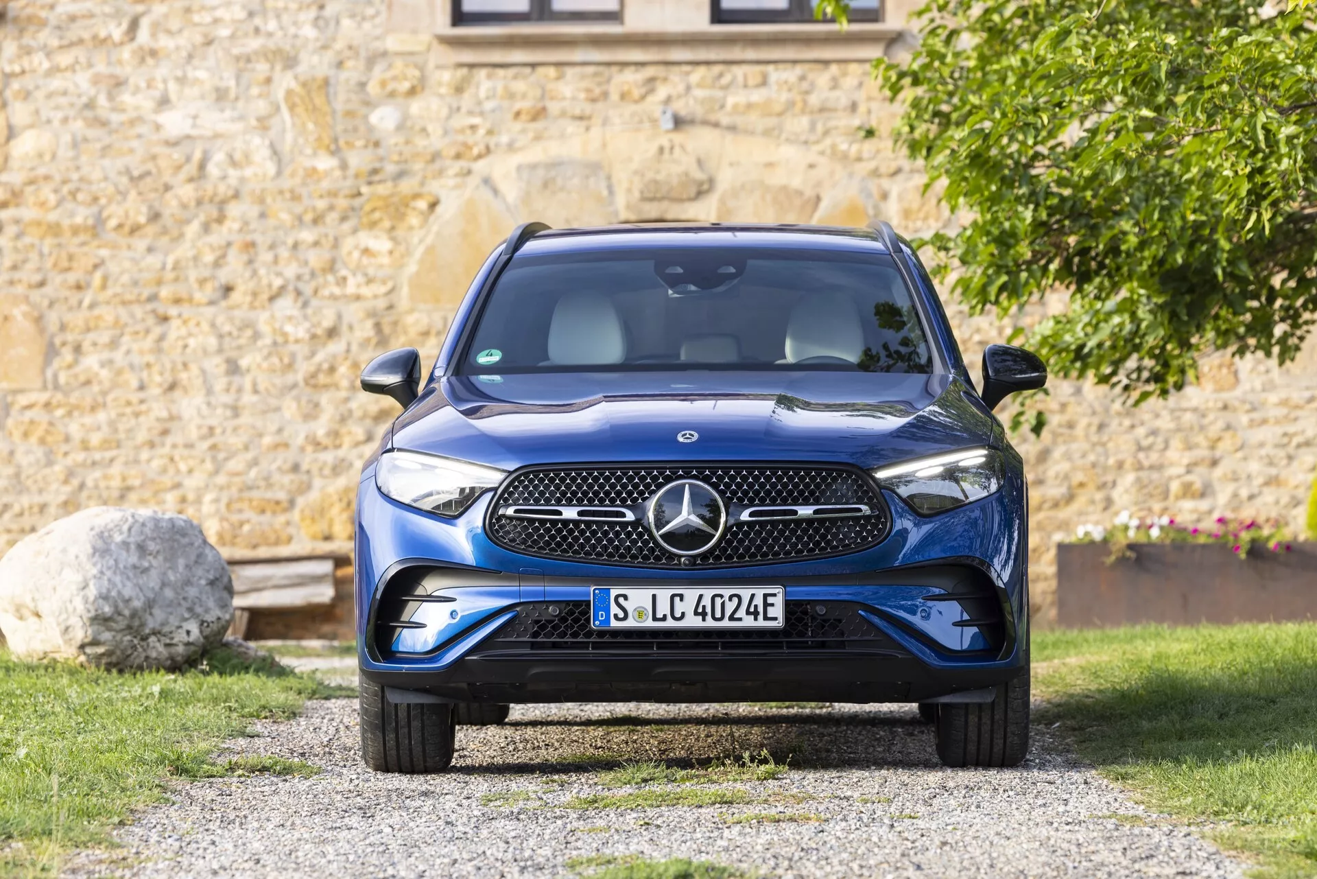 Frontal vy av en Mercedes GLC 2022 (254) som visar den framträdande frontgrillen, stötfångaren, motorhuven, strålkastarna och vindrutan. Bilen är placerad på en uppfart i en förortsmiljö med grönska och en del av en byggnad i bakgrunden. Grillen har en slät yta med en emblem som sannolikt innehåller sensorer eller en kamera. De eleganta strålkastarna verkar ha LED-teknologi. Den nedre stötfångaren inkluderar luftintag och parkeringssensorer. Den blå accenten på registreringsskylten indikerar att det är ett elfordon (EV) med specifika EV-funktioner. Den stora vindrutan ger god sikt och inkluderar troligen regnsensorer och en ljussensor. Sidospeglarna, även om de inte är helt synliga, är dörrmonterade och kan innehålla dödvinkelövervakning. Hjulens aerodynamiska design förbättrar räckviddseffektiviteten. Bilden är välbelyst med naturligt ljus och framhäver bilens detaljer och färg.