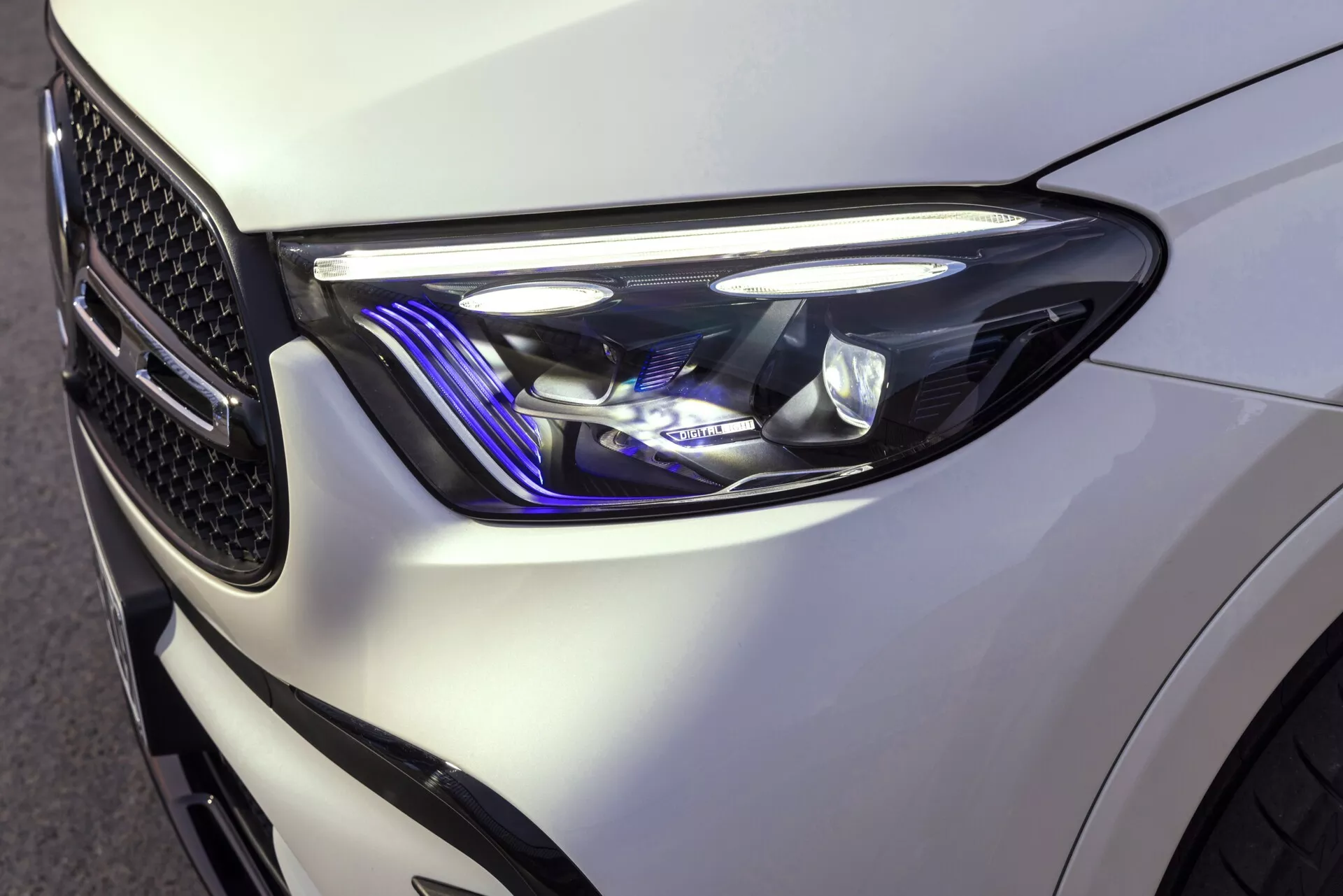 Närbild av den främre vänstra kvarten på en Mercedes GLC 2022, som visar den smidiga, moderna strålkastardesignen med skarpa, kantiga former och sofistikerade ljuselement, inklusive LED-dagljus och en blå nyans. Den glansvita färgen framhäver bilens väldefinierade linjer och det synliga honungskakemönstret på grillen speglar dess prestandainriktade design.