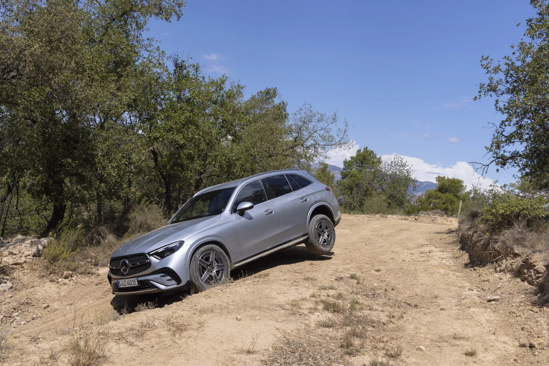 Bild av en Mercedes GLC 2022 SUV i en off-road-miljö tagen i en trekvartsvinkel från vänster framsida. Synliga funktioner inkluderar ett framträdande galler med centrerat emblem, LED-strålkastare med dagsljus, flerspoked fälgar, takräcken, skulpterad kaross, sidobackspeglar med integrerade blinkers och en förhöjd markfrigång som tyder på terrängkörningsförmåga. Fordonet är placerat på en grusväg, omgiven av gräs och träd, med naturligt solljus som belyser dess moderna design och lyxiga funktioner.