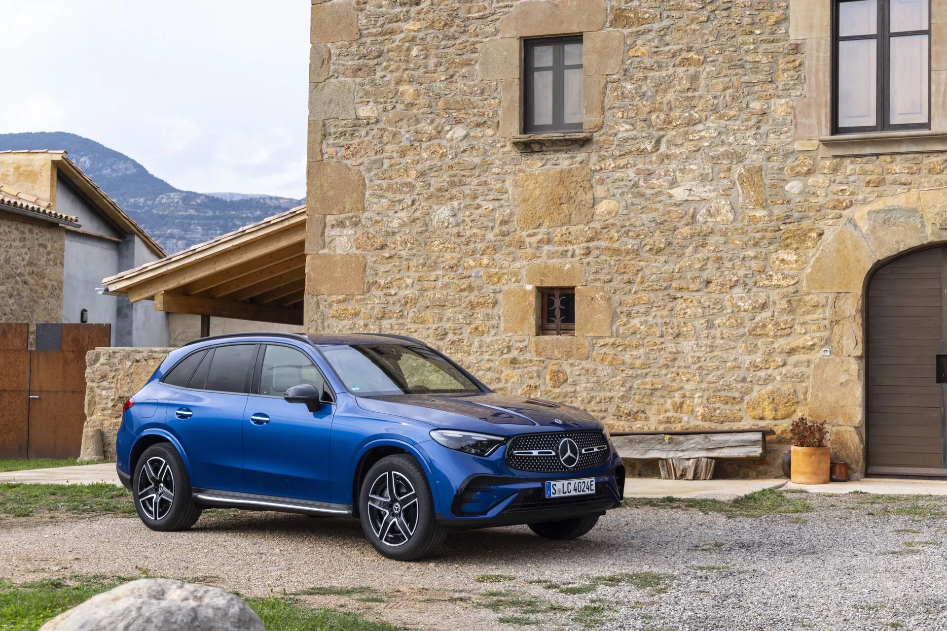 En modern blå Mercedes GLC 2022 SUV är framträdande visad utomhus, placerad i en lätt avig vinkel som framhäver den främre högra kvadranten och en del av sidprofilen. Bilen är prydd med en framträdande frontgrill med tillverkarens emblem, eleganta horisontella LED-strålkastare och stora flerspakiga aluminiumfälgar. Den står framför en gammal stenbyggnad, vilket ger en slående kontrast. Potentiella teknologiska funktioner som adaptiv farthållare, filhållningsassistent och panoramasoltak antyds av dess design. Bildens komposition erbjuder ett tydligt och naturligt perspektiv av fordonets lyxiga och högteknologiska attribut och fångar den detaljerade exteriören i en rustik miljö.