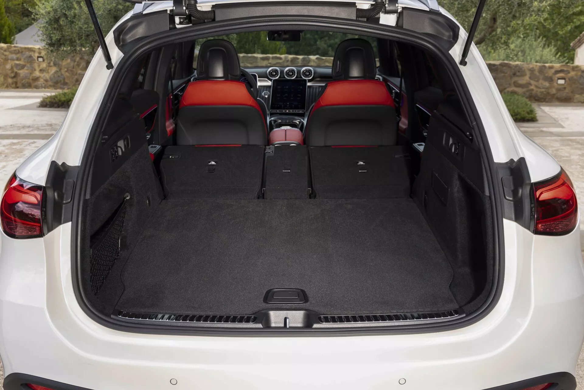 Bilden visar den bakre interiören av en Mercedes GLC 2022 (254) med bakluckan öppen. Vyn ger en tydlig bild av den rymliga, matta lastutrymmet, som har ett plant lastgolv och belyser flexibiliteten i utrymmet med de bakre sätena nerfällda i 60/40-konfiguration. Interiören har premium röda lädersäten, nackstöd med röda insatser och kontinuitet i design. Delvis synliga är instrumentbrädan och mittkonsolen, vilket antyder ytterligare funktioner som infotainment- och klimatsystem. Bakluckan antyder en användarvänlig design som kan inkludera teknik som handsfree eller motoriserad öppning. Ingen lastskydd är närvarande, vilket kan indikera att den är återdragbar eller borttagen.