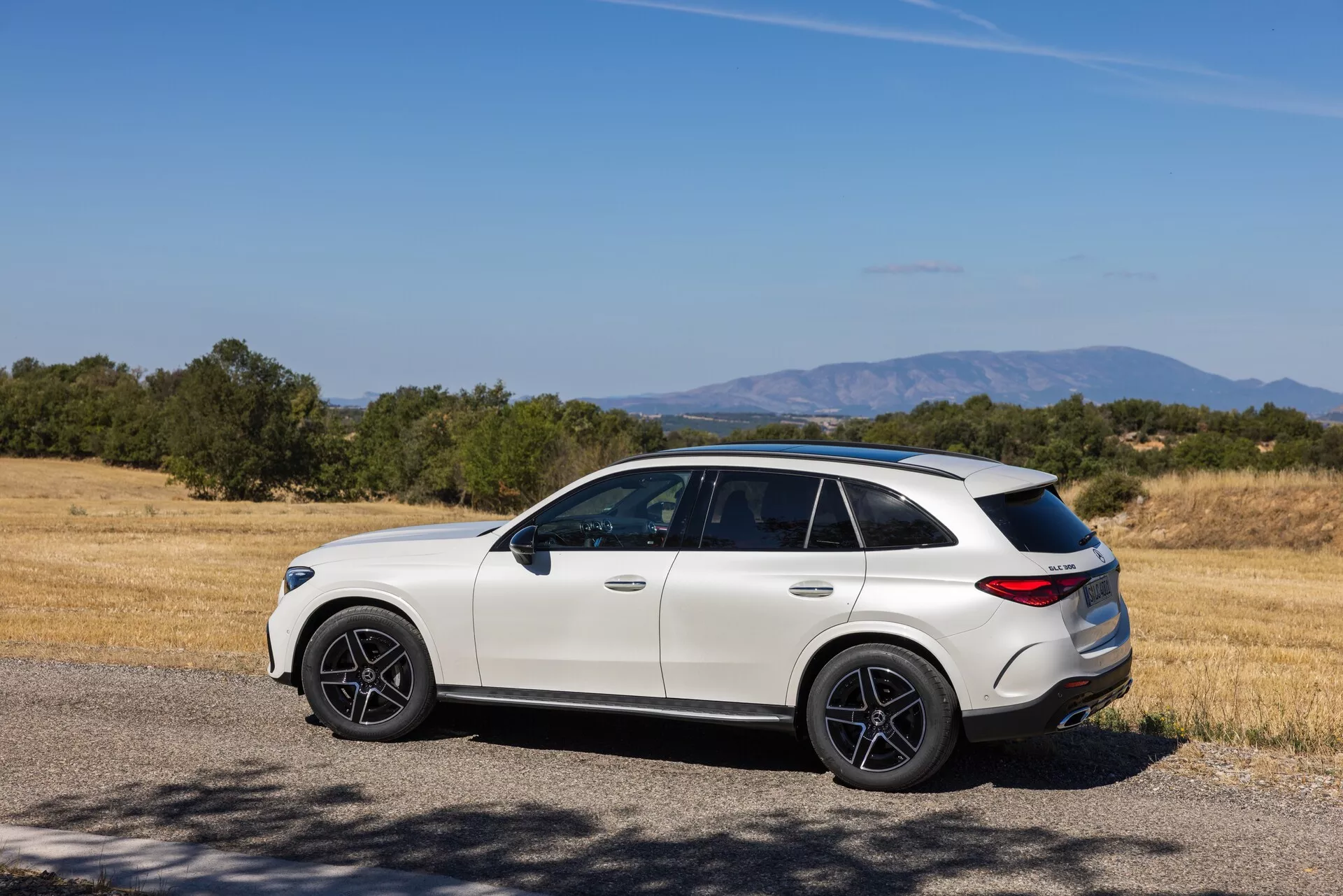 Modern lyxig crossover-SUV i lantlig miljö: trekvart bak-sidovy av en ljus Mercedes GLC 2022 (254) med sportiga mörka hjul, tonade fönster, sluttande taklinje och skulpterad bakre stötfångare, parkerad nära ett torrt gräsfält och träd under en klarblå himmel.