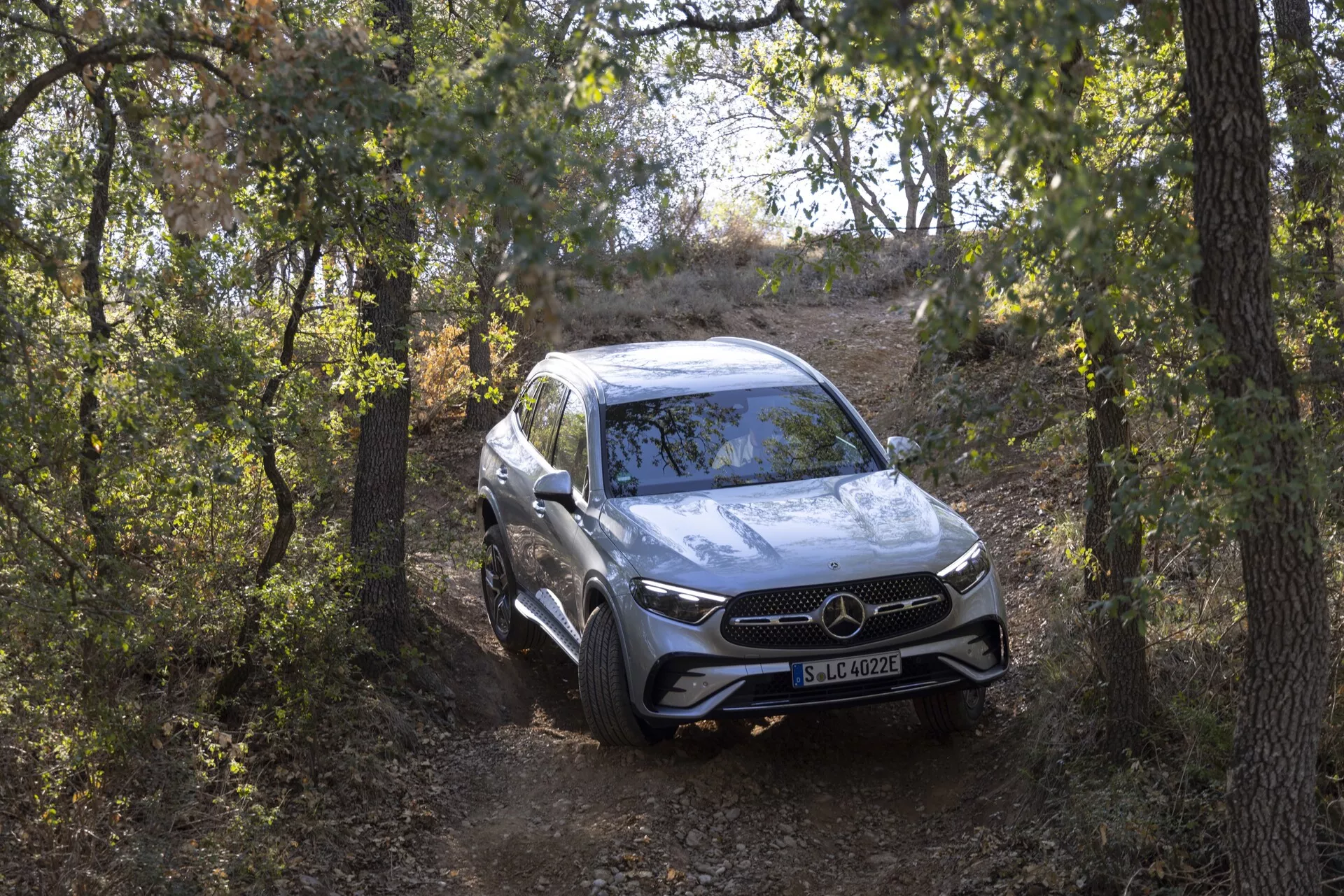 Full vy av Mercedes GLC 254 SUV från 2022 från ett främre trekvarts perspektiv närmar sig kameran på en naturlig stig omgiven av träd. Den något höga vinkeln betonar dess terrängförmåga, körhöjd och fjädringsinställning. Lyxbilen har en framträdande frontgrill med ett stort emblem, LED-strålkastare, parkeringssensorer och aerodynamiska konturerade linjer på huven. Den silverfärgade lacken speglar omgivningen medan lättmetallhjul och mångsidiga däckskvalpar bidrar till ett sportigt och elegant utseende.