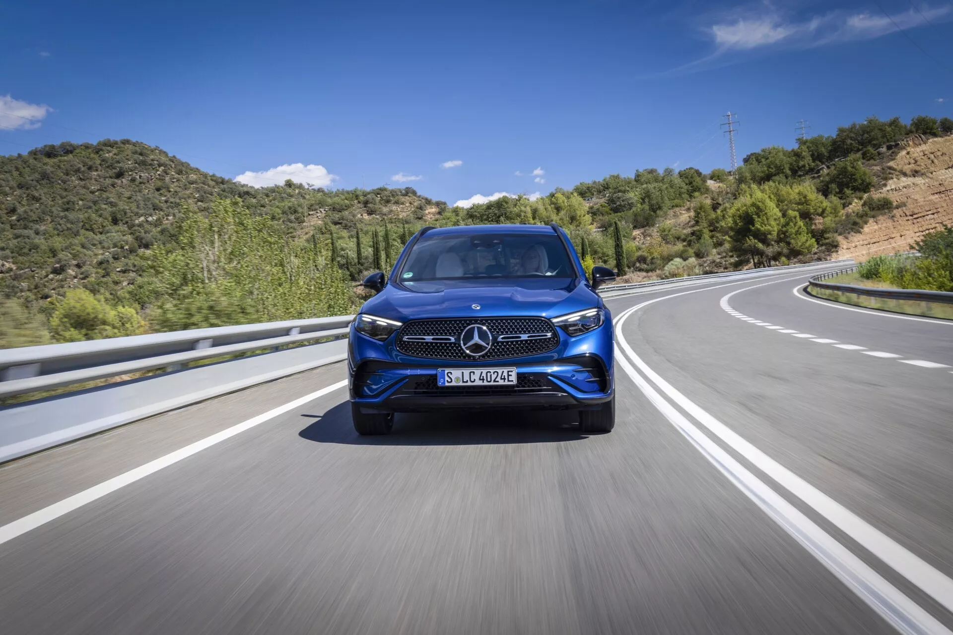 En låg vinkel, frontal vy av en modern Mercedes GLC 2022 som kör på en motorväg eller landsväg. Den här bilden framhäver fordonets distinkta grill, strålkastarens design, frontstötfångarens stil, stora lättmetallfälgar och potentiella avancerade förarassistanssystem (ADAS). Belysningen är ljus och naturlig med en bakgrund av blå himmel och gröna kullar, vilket antyder dess rörelse på asfalt med delade filmarkeringar.