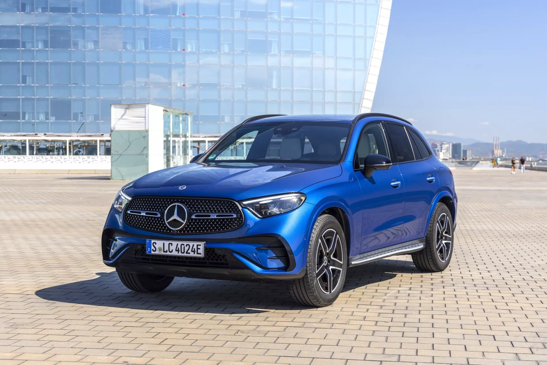 Trekvartsvy framifrån av en blå Mercedes-Benz GLC SUV 2022 som visar den karakteristiska Mercedes-grillen med emblemet, eleganta horisontella ribbor, integrerad stötfångare med dimljus och luftintag, skarpa LED-strålkastare, konturerade motorhuvslinjer, sportiga mångekrade lättmetallfälgar, kaross-färgade handtag och speglar, tonade fönster, soltak och möjliga avancerade förarassistanssystem. Fotograferad i ljus utomhusmiljö med naturligt solljus och klarblå himmel.