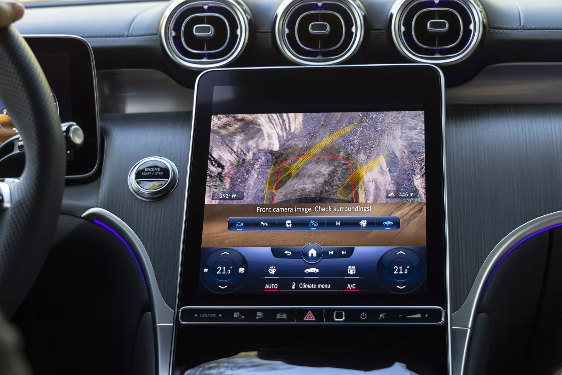 Närbild av interiören i en Mercedes GLC 2022 som visar mittkonsolen från en passagerares perspektiv. Fokus ligger på den vertikalt orienterade pekskärmen som visar en vy från frontkameran med parkeringslinjer, klimatanläggningsknappar som visar tvåzons temperaturinställningar på 21°C, runda luftventiler ovanför skärmen och en start/stopp-knapp för motorn. Bilden framhäver avancerade infotainmentsystem, parkeringsassistans, tvåzons klimatanläggning och material av premiumkvalitet i interiören.
