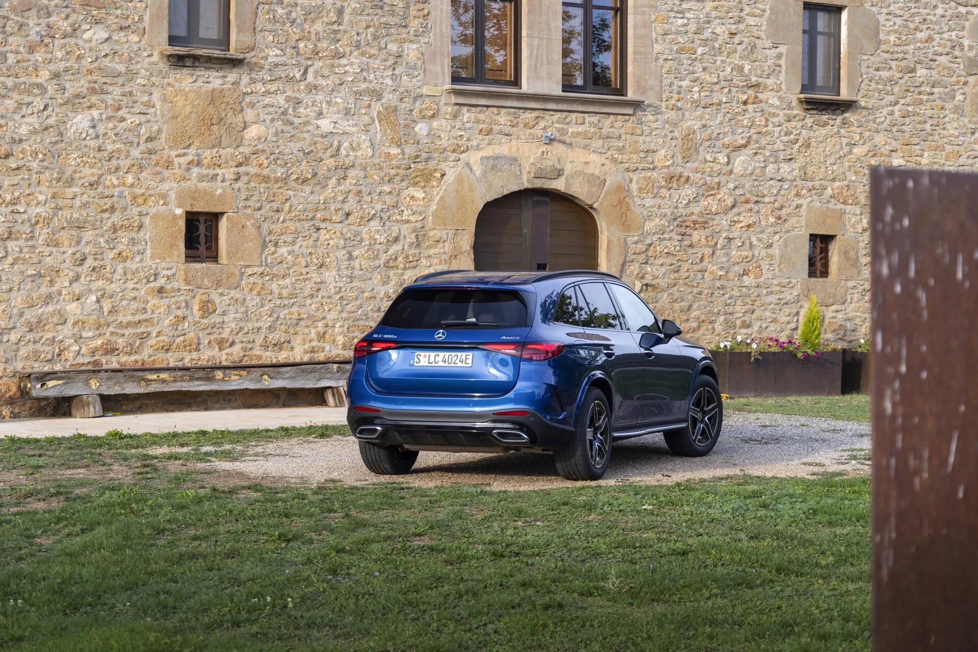 Modern SUV parkerad på en grusväg sedd från trekvart bakifrån. Bakljusen har modern LED-teknik sömlöst integrerad i designen. Bakrutan är lätt framåtlutad och bidrar till en aerodynamisk profil, ackompanjerad av en takspoiler. Bakre stötfångaren har inbyggda sensorer eller reflektorer som antyder ett parkeringsassistanssystem och visar dubbla avgasutblås inbyggda i den nedre delen av stötfångaren. SUV:n har stora fälgar i flera ekrar och en ren, rik blå yta som reflekterar ett väl underhållet utseende mot bakgrunden av en rustik stenbyggnad.