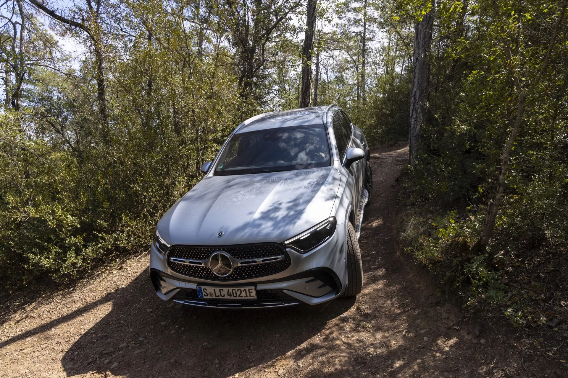 Modern lyxbil, Mercedes-Benz GLC 2022, hög främre vinkelperspektiv som visar framgrill, stötfångare, motorhuv, vänster sida inklusive förardörr och sidospegel, elegant samtida design, framträdande Mercedes-Benz treuddsembem, detaljerade LED-strålkastare, flerekrade aluminiumhjul, potentiellt EQ-märke som indikerar elektrisk eller hybridmodell, sannolikt ADAS-funktioner, placerad på smal grusväg omgiven av vegetation, silver eller ljusgrå metalliclack som framhäver premiumkvalité.