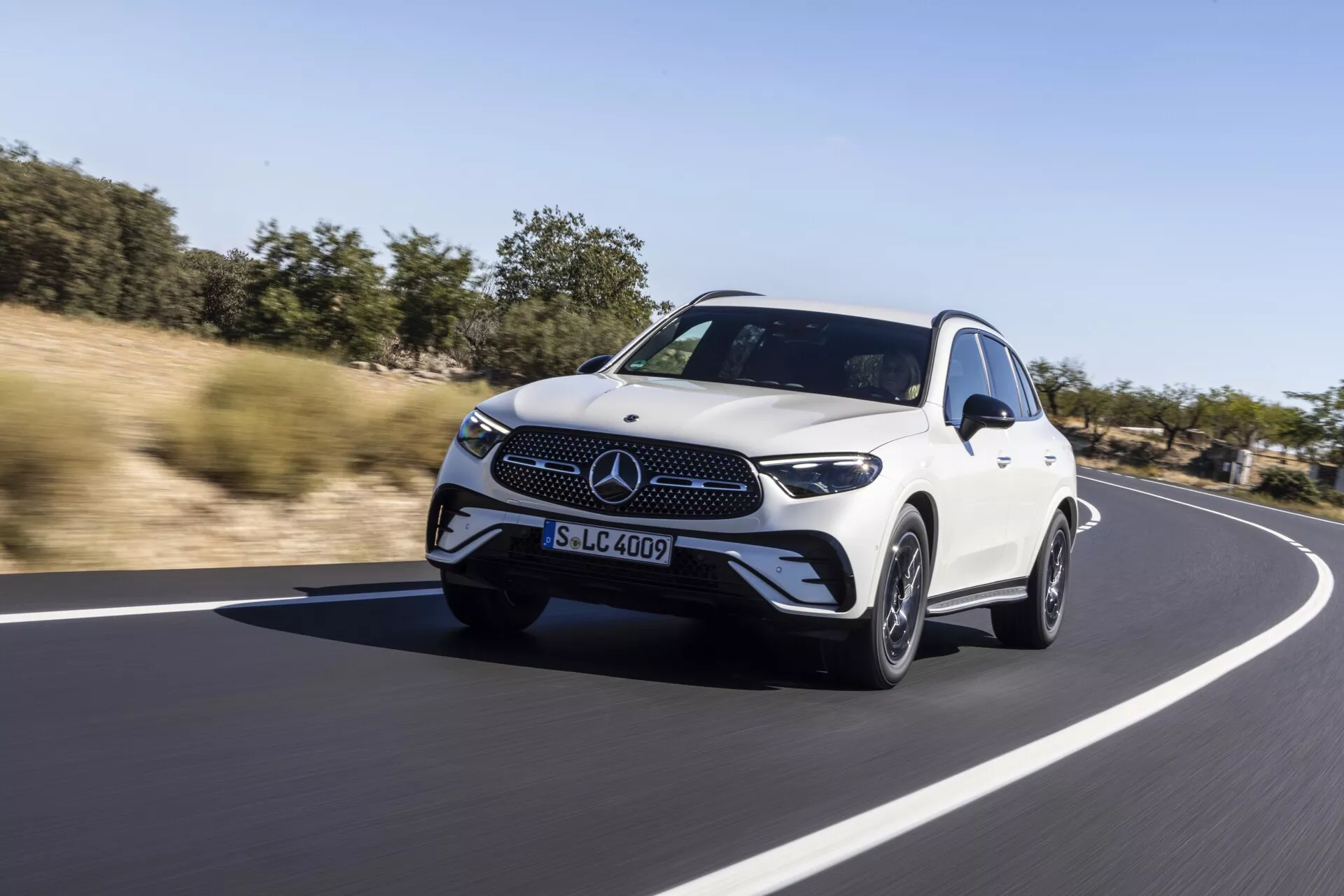 Lyxig vit Mercedes GLC 2022 crossover SUV som kör på en jämn asfaltväg, naturligt landskap i bakgrunden under klar dagtidshimmel. Lågvinklad dynamisk bild som fångar en trekvartsvy framifrån med LED-strålkastare, framstående frontgrill med emblem, aerodynamisk design och stiliga lättmetallfälgar, betonar känslan av hastighet och rörelse med lätt rörelseoskärpa i bakgrunden.