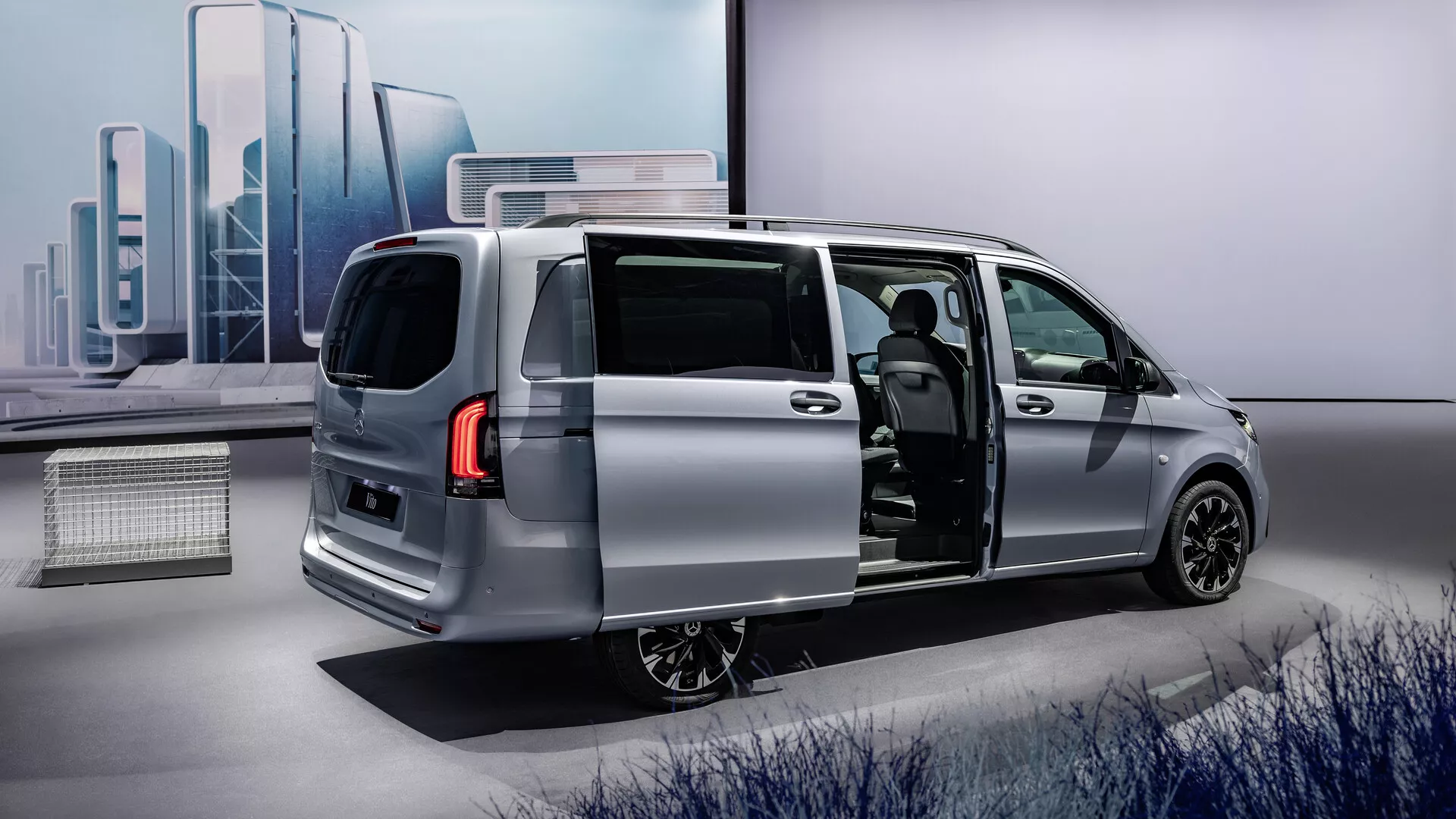 2024 Mercedes-Benz Vito (447 facelift) silver minivan med öppen skjutdörr på sidan som visar den eleganta moderna designen från ett trekvartsbakperspektiv, framhäver fordonets aerodynamiska profil, LED-bakljus, legeringshjul och en titt in i den avancerade interiören med potentiellt premium sätesmaterial och en modern instrumentbräda.