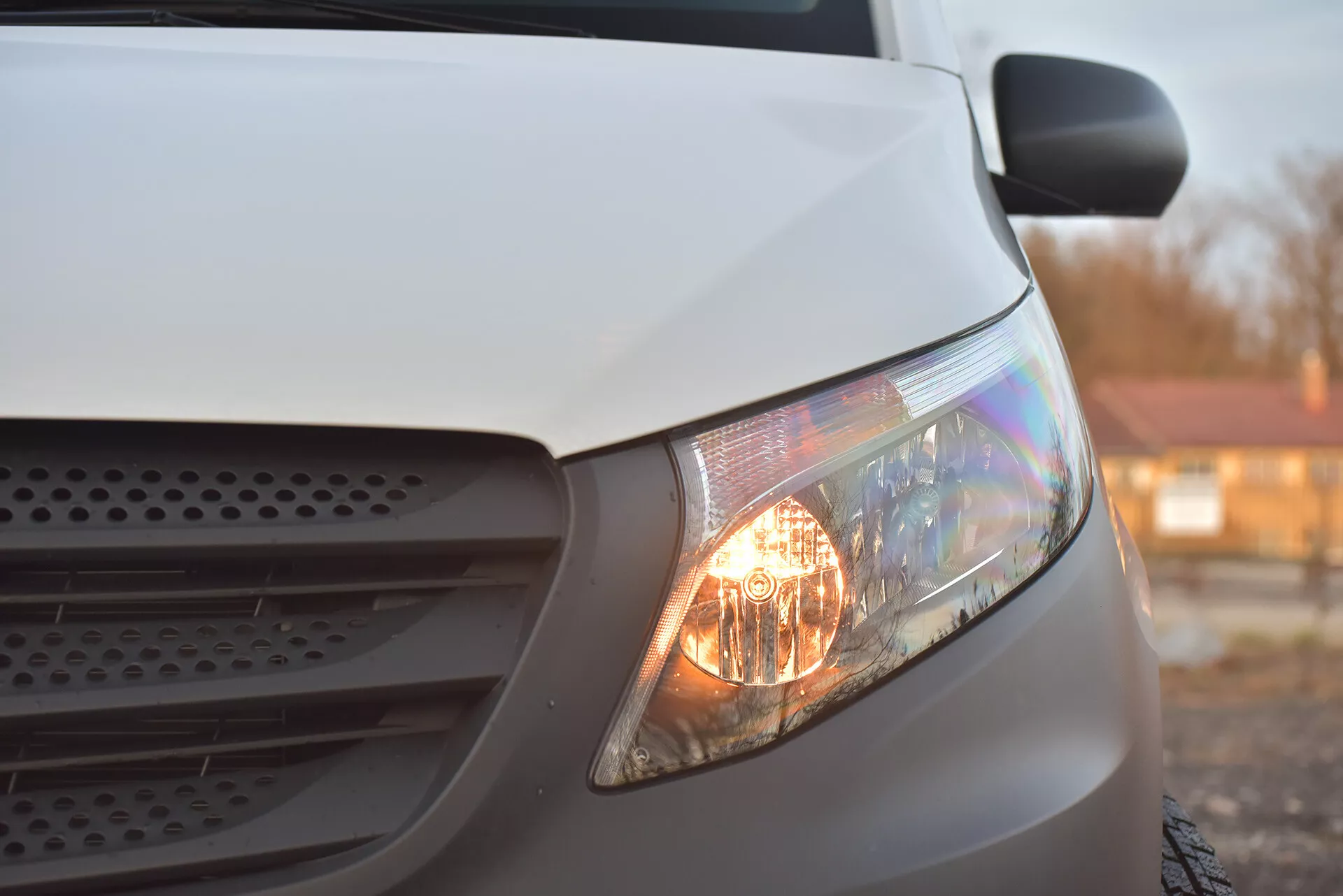 Närbild på sidovy av en vit Mercedes-Benz Vito Tourer från 2014 med fokus på den belysta strålkastaren, en del av grillen och motorhuvens sektion, framhäver dess halogenstrålkastare och aerodynamiska design under mjukt ljus.