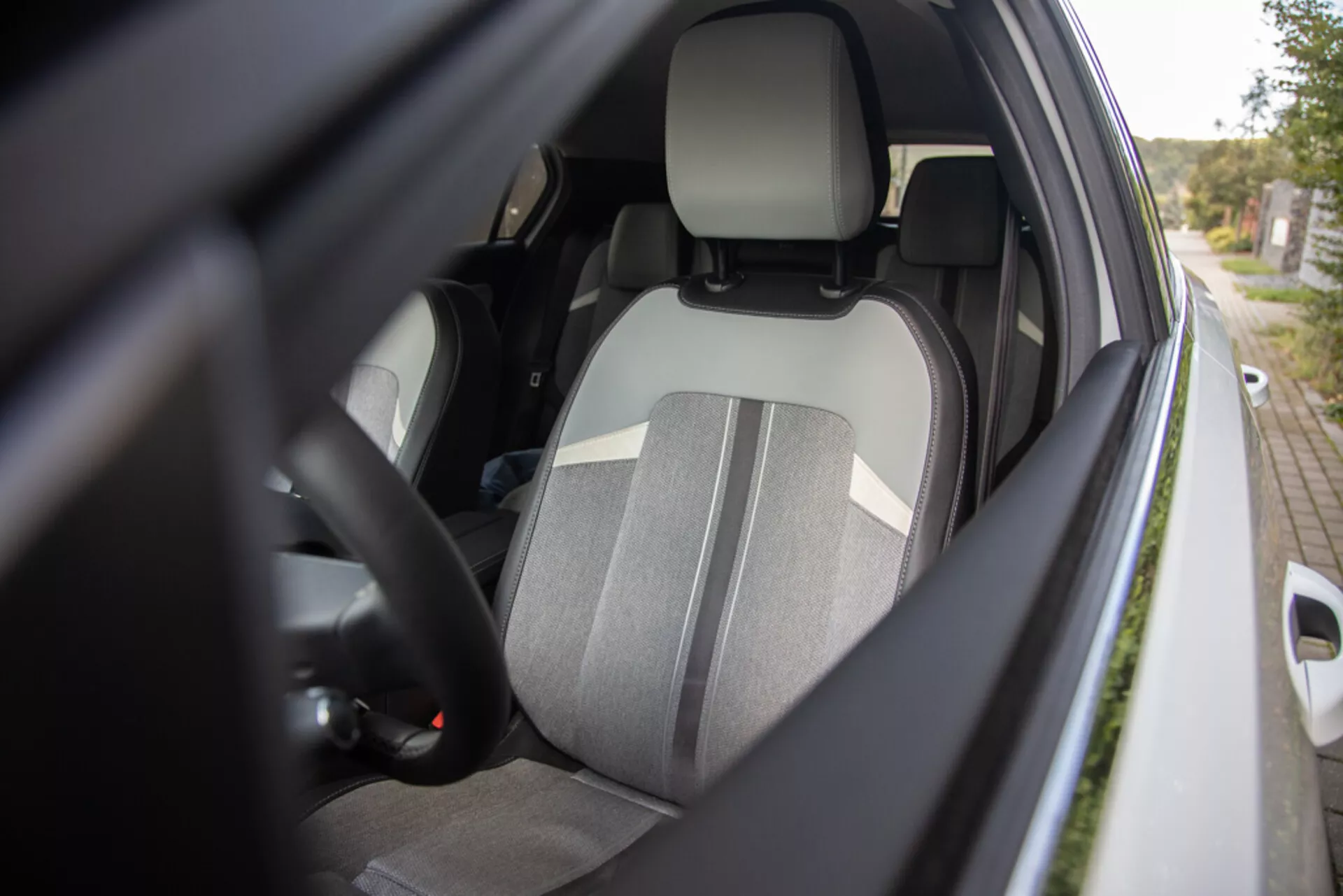 Närbild av förarsidans interiör i Opel Astra 2022 från en yttre sidovinkel som visar förarsätet med tvåtonat tyg, integrerat nackstöd, del av ratten med kontroller, glimt av instrumentbrädan, fastspänt säkerhetsbälte, dörrinteriör med potentiella elektriska fönsterkontroller och karossfärgat sidospiegelhus med fokus på ergonomi, säkerhet och bekvämlighetsfunktioner.