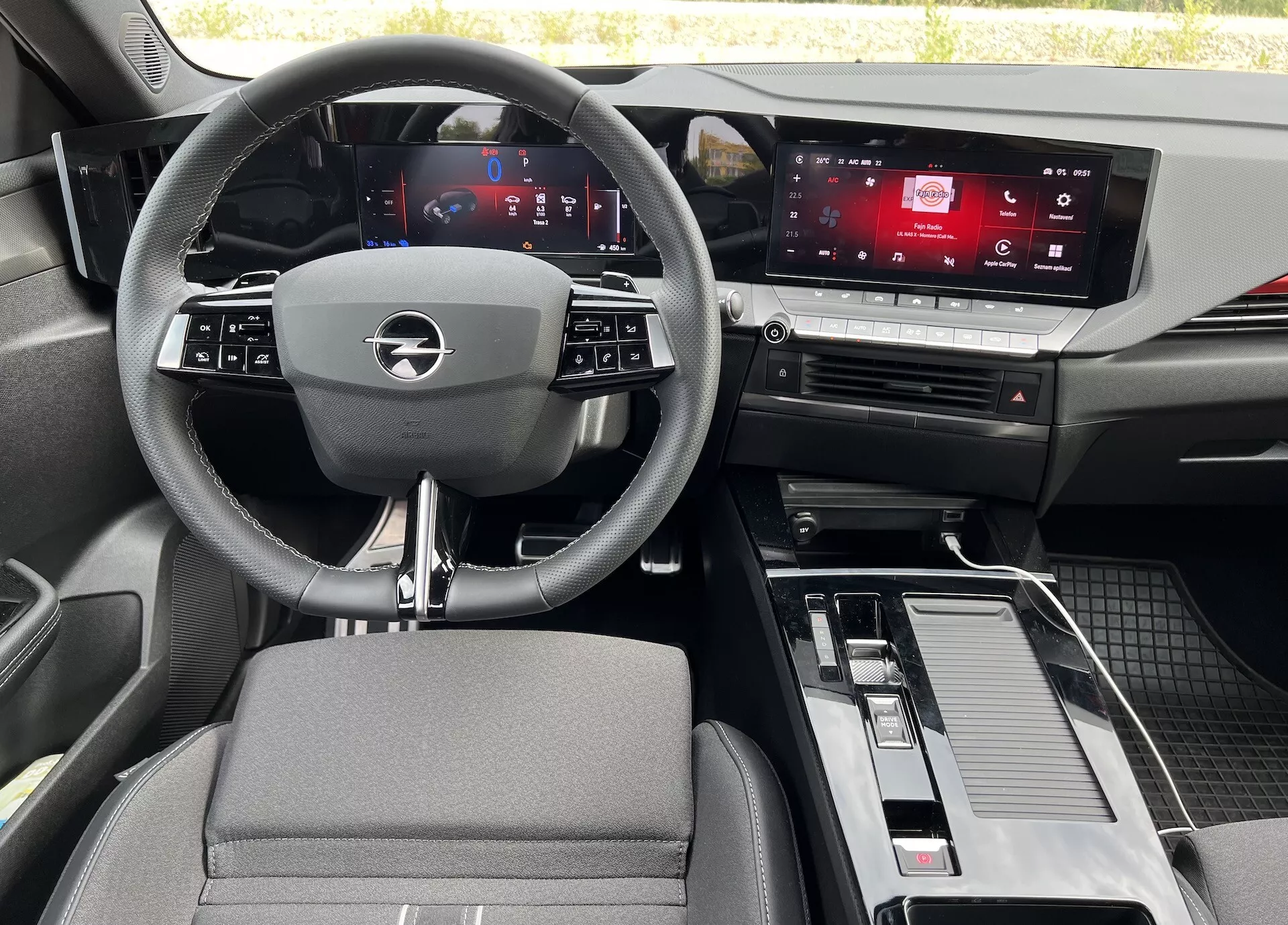 Interiörvy av Opel Astra 2022 från baksätet, visar en trespokig ratt med integrerade kontroller, digital instrumentkluster, pekskärm för infotainmentsystem, mittkonsol med växelspak och förvaring samt en blandning av tyg- och metall-effektmaterial