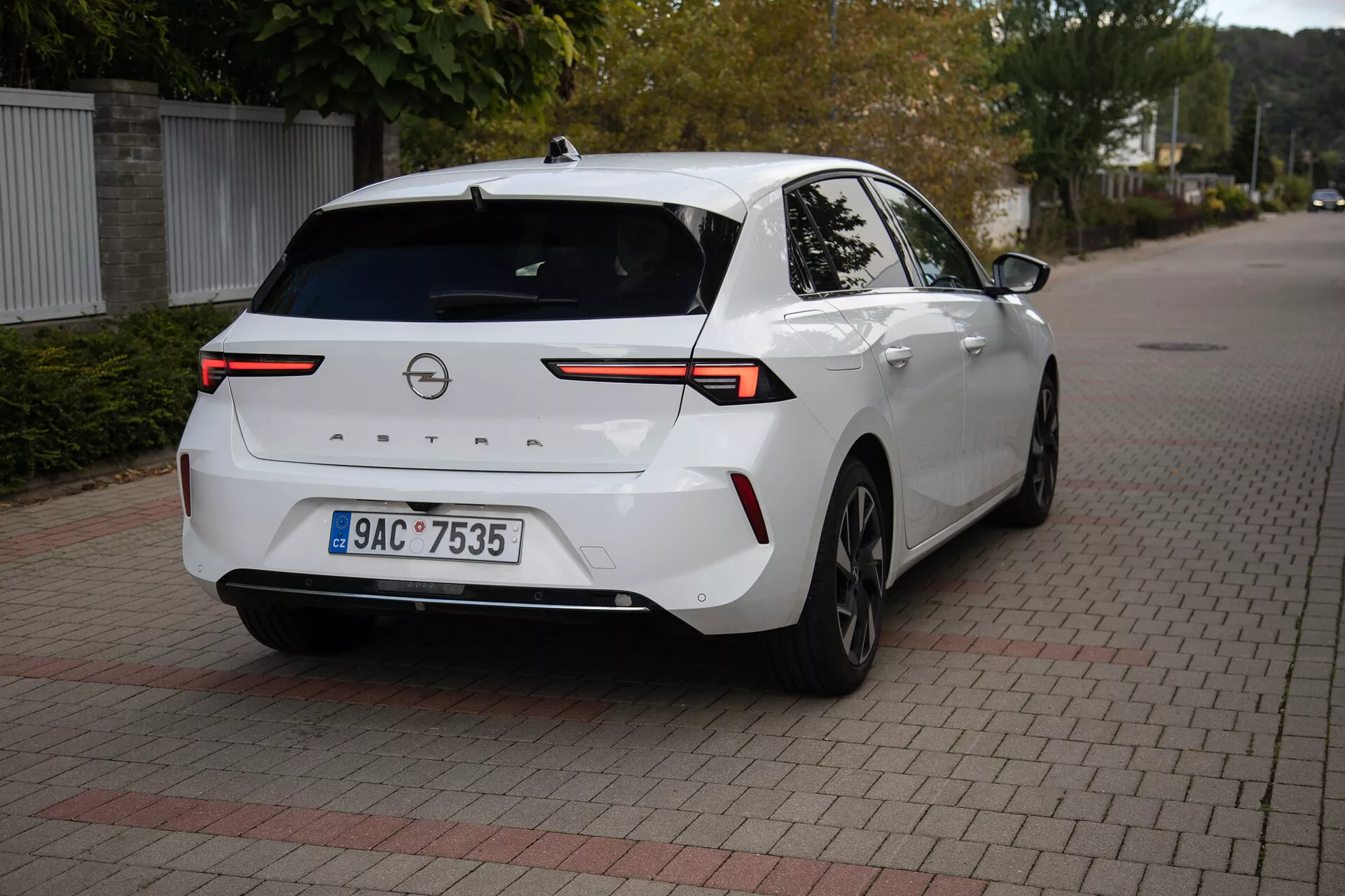 Modern sedan Opel Astra 2022 i vitt, parkerad på yta av sammanlåsande tegelstenar, visar bakifrån tre kvartals vinkel med fokus på högteknologiska LED-bakljus, backkamera, parkeringssensorer, delvis sidoprofil inklusive höger bakhjul och dörr, förbunden med en horisontell ljuslist över bagageluckans lock, utrustad med hajfensantenn för förbättrad anslutning, lämplig för att illustrera bakre fordonets design och modern fordons teknologi