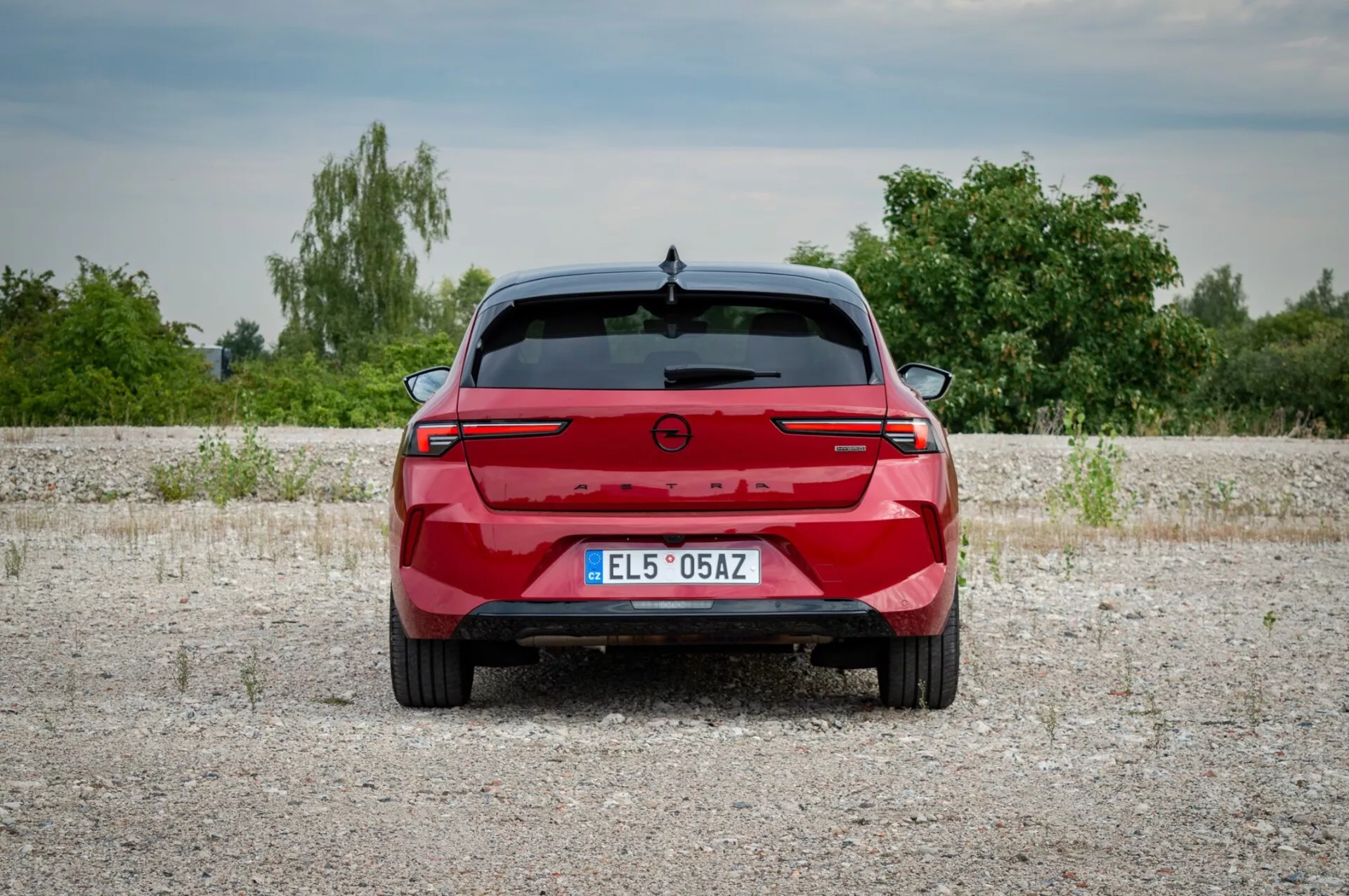 Opel Astra 2022 fotograferad bakifrån på en grusyta som visar bakljus med en anslutande ljuslist, parkeringssensorer och avsaknad av avgasportar som antyder en potentiell elektrisk eller hybridmodell, mot en bakgrund av vegetation och lätt molnig himmel.