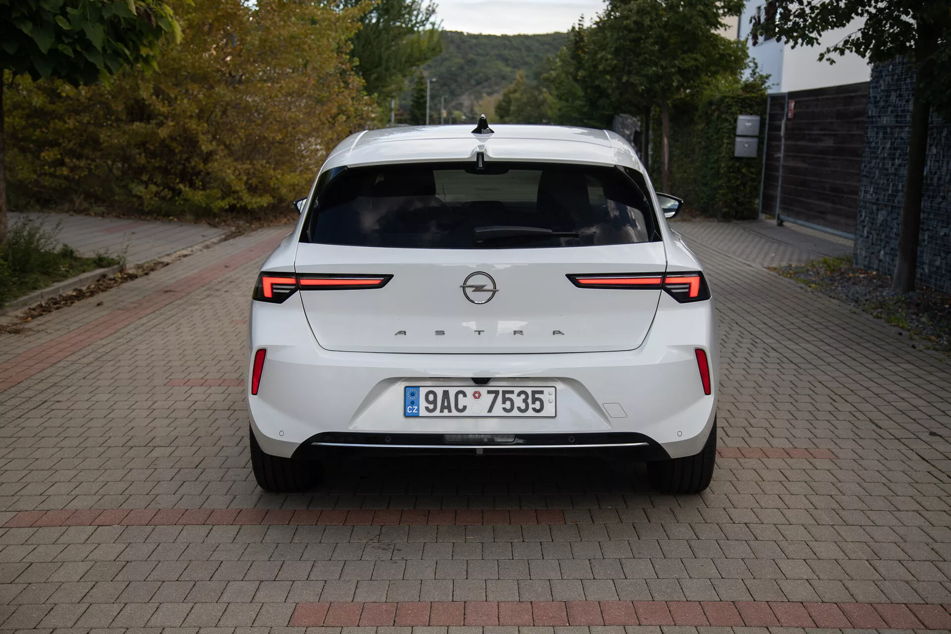 Bakifrån vy av en vit Opel Astra 2022 som visar LED-baklampor, parkeringssensorer/kamera bak, märkesmärke, registreringsskylt, bakspoiler, högt monterat stoppljus, hajfensantenn och ett rent lackfinish, parkerad på en tegelmönstrad yta i dagsljus.