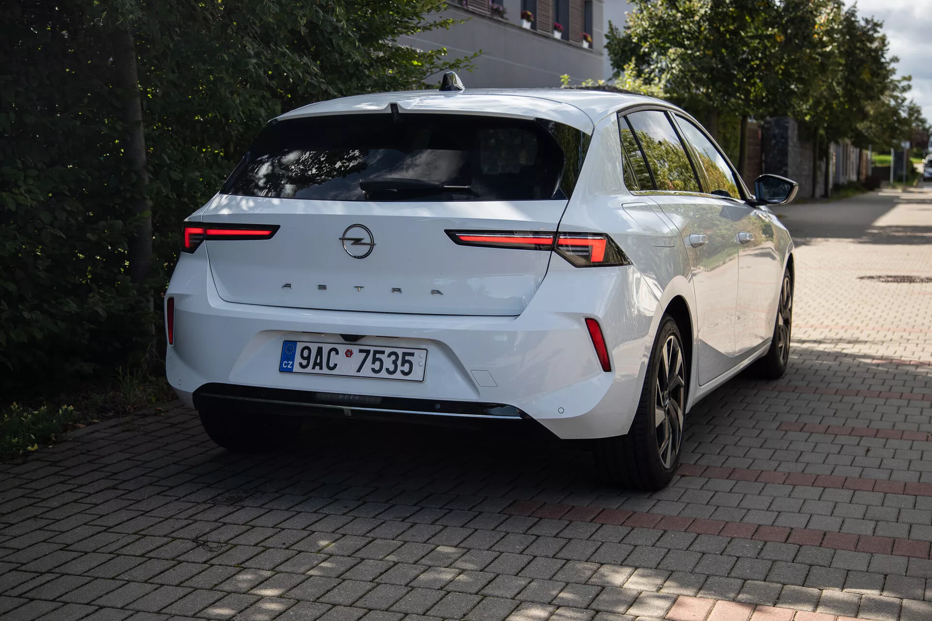 Vit Opel Astra hatchback 2022 fotograferad från en bakre tre fjärdedels vinkel som framhäver dess långsträckta horisontellt orienterade bakljus, böjt baklucka med integrerad spoiler och extra bromsljus, tillverkarens logotyp och modellbeteckning på bakluckan, centralt placerat registreringsskylt, glimt av bakre stötfångaren, flerspakiga legeringshjul, snygg sidoprofil med en märkbar karaktärslinje, bakre fönster som smalnar av mot bilens baksida, bakre fönster med torkare, och en hajfen antenn, parkerad på ett tegelbelagt område med träd och byggnader i bakgrunden.
