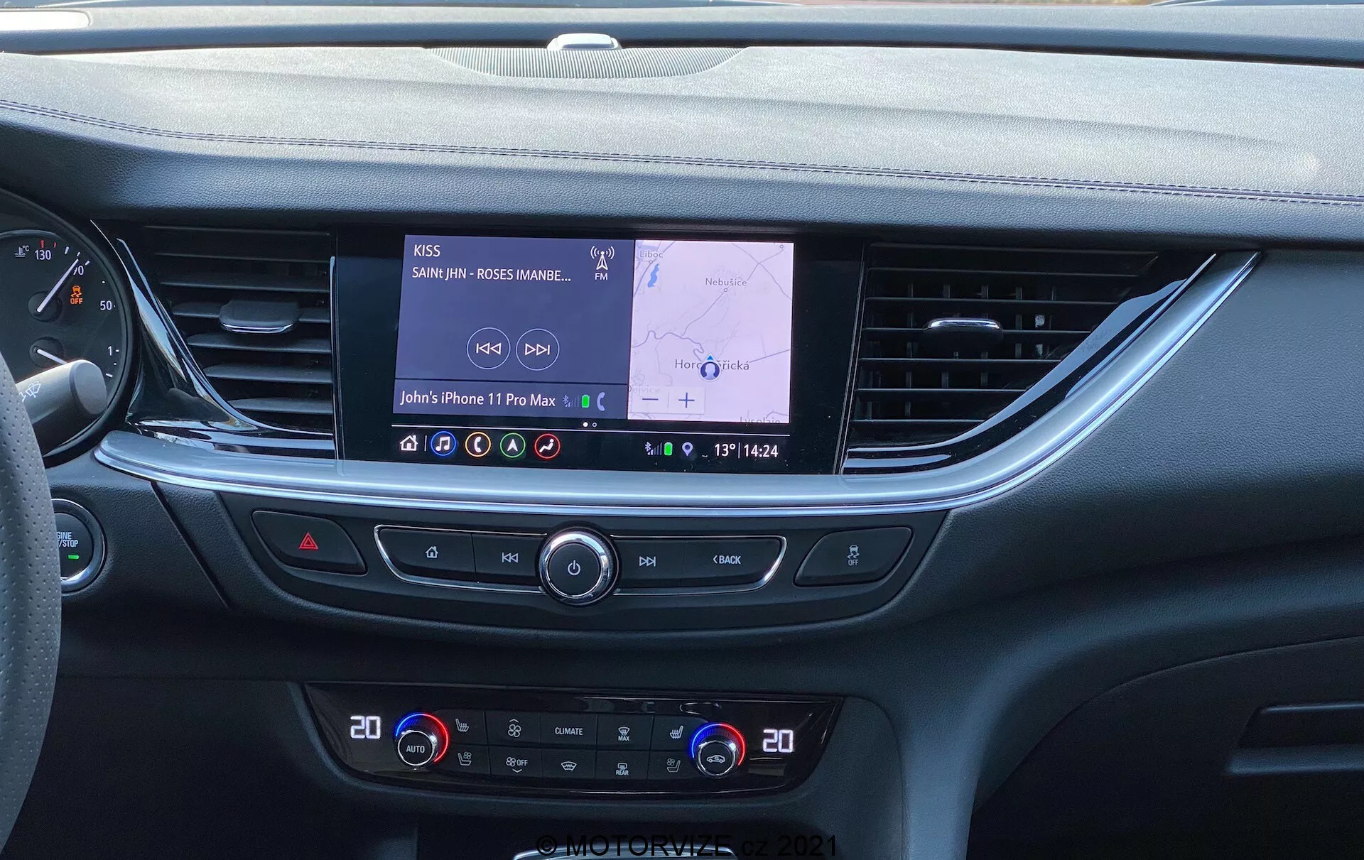 Interiören av en modern 2020 Opel Insignia GSI Grand Sport, som visar mittkonsolen och infotainmentsystemet från en ögonhöjdperspektiv. En stor rektangulär pekskärm med en aktiv GPS-rutt och Bluetooth-ljudanslutning dominerar konsolen, omgiven av klimatanläggningsrattar och knappar. Den eleganta instrumentpanelens design har mjuka material, horisontella linjer och sträcker sig in i dörrpanelerna. Under klimatanläggningskontrollerna är ytterligare knappar synliga för funktioner som bakrutedefrost, recirkulation av kabinluft och start/stopp-system, tillsammans med en förvaringsfack eller trådlös laddplatta längst ner.