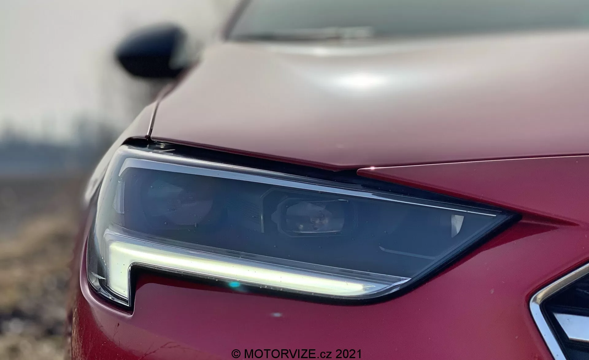 Närbild på Opel Insignia 2020 strålkastarenhet, fokuserad på fordonets vänstra framkvartspanel. Den moderna designstrålkastaren har klara linser med LED- eller HID-teknik, och producerar en skarp, vinklad ljussignatur. Fotografiet framhäver strålkastarens skarpa design och inkluderar kromaccent, en delvis vy av frontgrillen och en metalliskt målad motorhuv, uppnådd med ett grunt skärpedjup och bokeh-effekt för en konstnärlig touch.