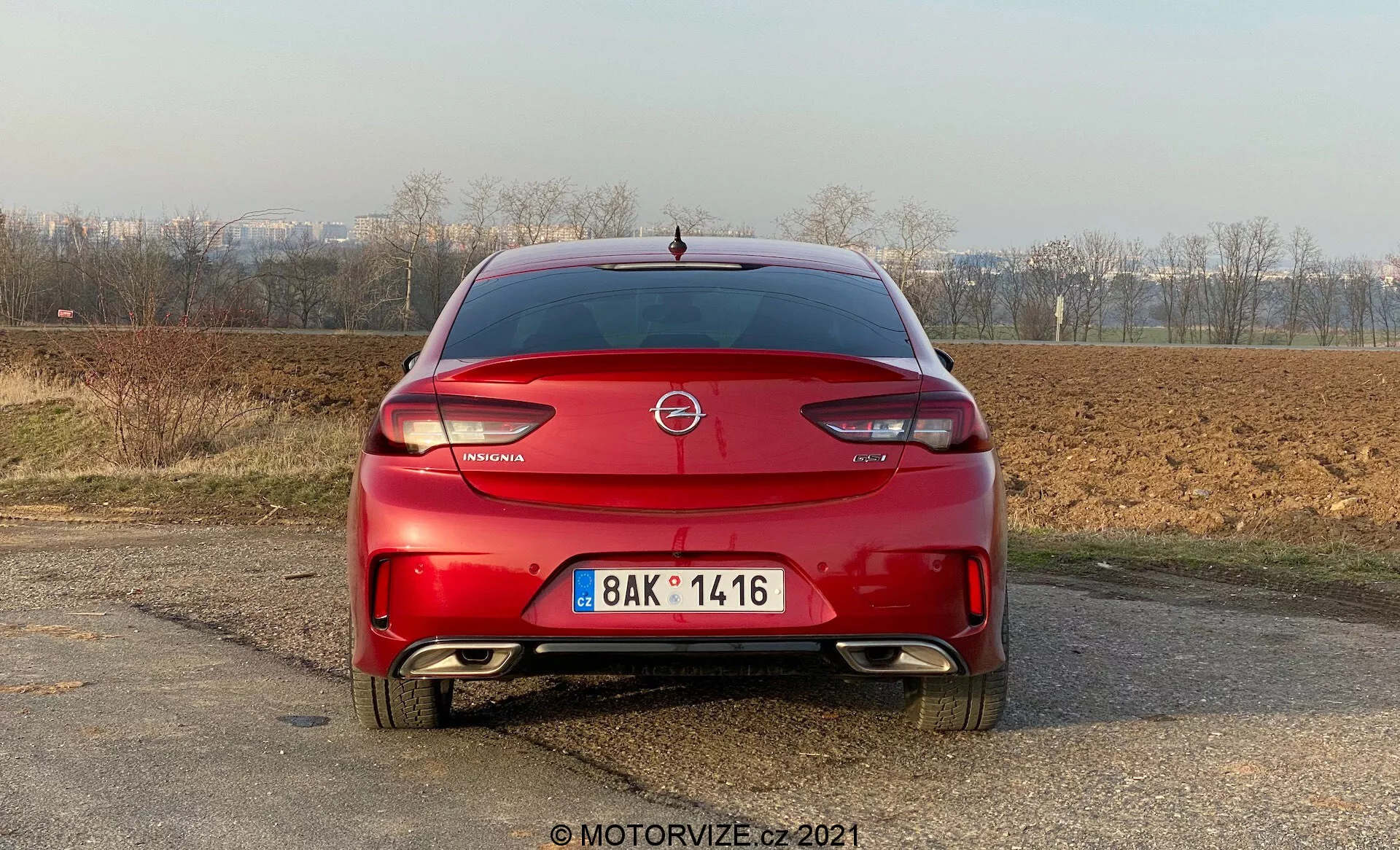 Bakifrånvy av en röd Opel Insignia GSi Grand Sport 2020 parkerad på en grusig yta med ett naturskönt, möjligen landsbygdsliknande landskap i bakgrunden. Fotot, taget på eftermiddagen i naturligt ljus, visar bilens emblem tydligt på bagageluckan, med modellnamnet under. Högt monterat bromsljus syns under bakrutan. Baklyktorna har en svepande design runt sidorna på fordonet, vilket är kännetecknande för modern design. Bakre stötfångaren har reflexer och eventuellt parkeringssensorer, med ett dubbelt avgasystem som indikerar en sportig design. Registreringsskylten med ett synligt registreringsnummer är centralt placerad på stötfångaren. En liten spoiler finns på kanten av bagageluckan.