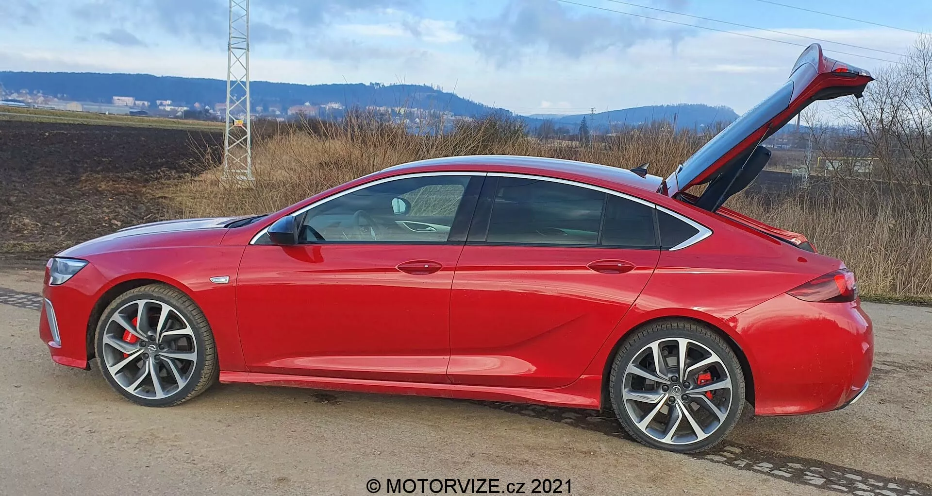 Ljus röd, modern 2020 Opel Insignia-GSI-Grand Sport sedan i sidovy, riktad höger, på en vägsida med naturlandskap; funktioner inkluderar aerodynamiska linjer, sluttande taklinje, öppen bagagelucka med automatisk öppningsfunktion, stora fler-ekrade lättmetallfälgar, karossfärgade sidospeglar med integrerade blinkers, synliga skivbromsar med ok, något tonade fönster, metallisk färg, skarpa karaktärslinjer och avancerad dörröppningsteknik.