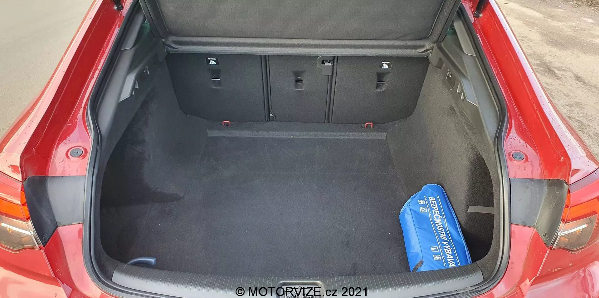 Högvinklig bild av det öppna bagageutrymmet på en röd 2020 Opel Insignia GSi Grand Sport, sett bakifrån. Det rymliga bagageutrymmet har ett mörkt, mattbelagt golv och 60/40 delbara fällbara baksäten i upprätt läge. Konturer och urtag på sidorna rymmer strukturella element, vilket maximerar det användbara utrymmet. Ett enda blått objekt på höger sida fungerar som referenspunkt för storlek.