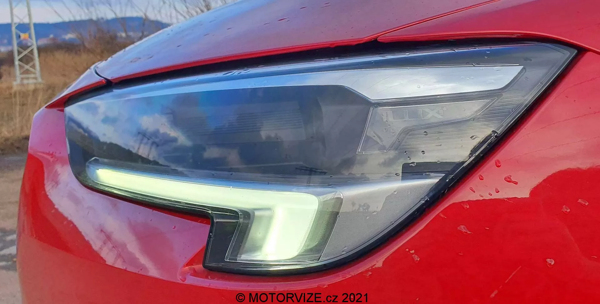 Närbild av Opel Insignia 2020 GSi Grand Sports vänstra främre strålkastarenhet, som visar en modern, elegant design med LED-varselljus som böjer sig uppåt längs ytterkanten. Den livfulla röda huven, skärmen och den klara strålkastarhöljet framhäver bilens aerodynamiska design och moderna belysningsteknik. Små vattenfläckar och lite smuts tyder på att bilen används regelbundet.