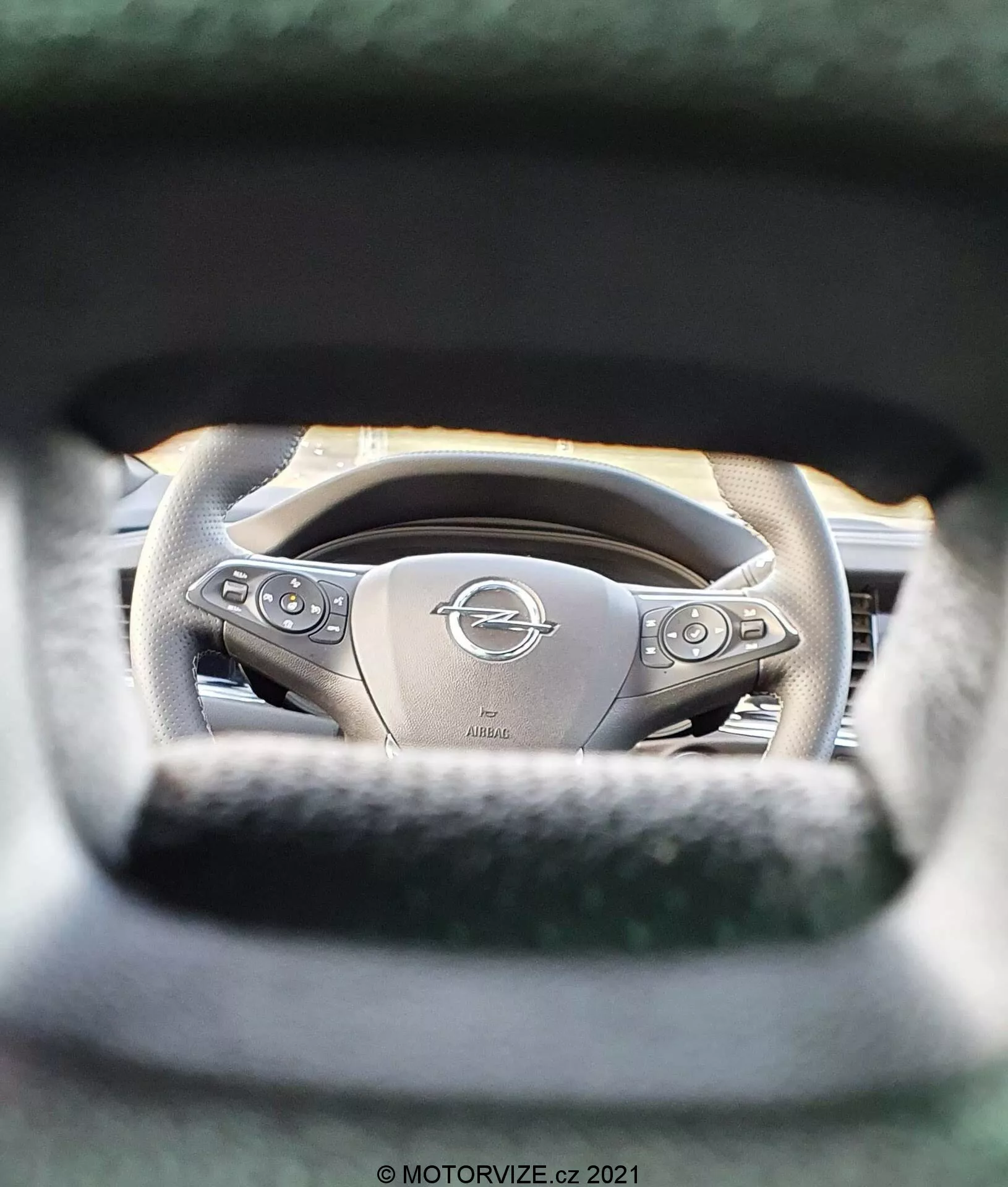 Interiör av Opel Insignia 2020 (Insignia GSi Grand Sport) med en detaljerad vy av ratten från förarsättets perspektiv. Fokus ligger på den Opel-emblemcentrerade, ergonomiskt designade läderklädda ratten med multifunktionella knappar. Det smala skärpedjupet framhäver ratten medan det omgivande området, inklusive instrumentpanelen, är suddig. Märkbara funktioner inkluderar metalliska accenter, en möjlig uppvärmningsfunktion och en gravering 'AIRBAG', som betonar både lyx och säkerhet.