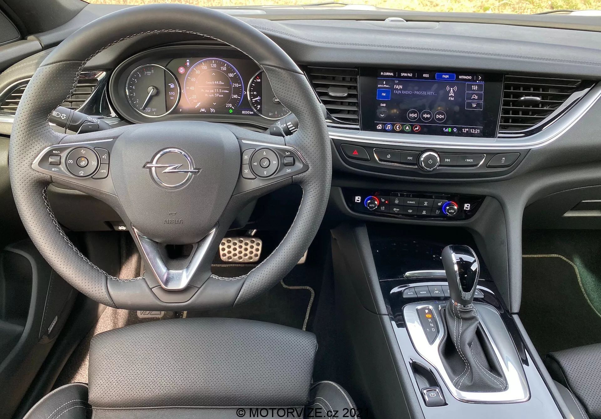 Interiörsbild av en Opel Insignia GSi Grand Sport 2020 från baksätet, som visar en multifunktionsratt med Opel-logotyp, delvis synligt analogt och digitalt instrumentkluster, framträdande infotainmentskärm med pekfunktion, klimatkontrollpanel, automatisk växelspak med lädermanchett och silverdetaljer, mugghållare, mittkonsol med förvaringsutrymme och en mörk, samtida interiördesign med metalliska inslag, vilket tyder på ett modernt och möjligen premiumfordon.