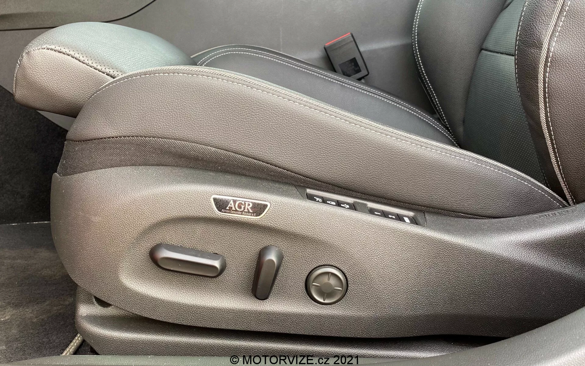Närbild på sidan av framsätet i en Opel Insignia GSi Grand Sport 2020, med fokus på sätesjusteringskontroller. Bilden visar mörkgrå eller svart klädsel, sidoknappar för fram- och bakpositionering, vertikala knappar för ryggstödsjustering, ett runt vred för svankstöd, en 'AGR'-logga och en spak som troligen reglerar säteshöjden. © MOTORVIZE.cz 2021 vattenstämpel.