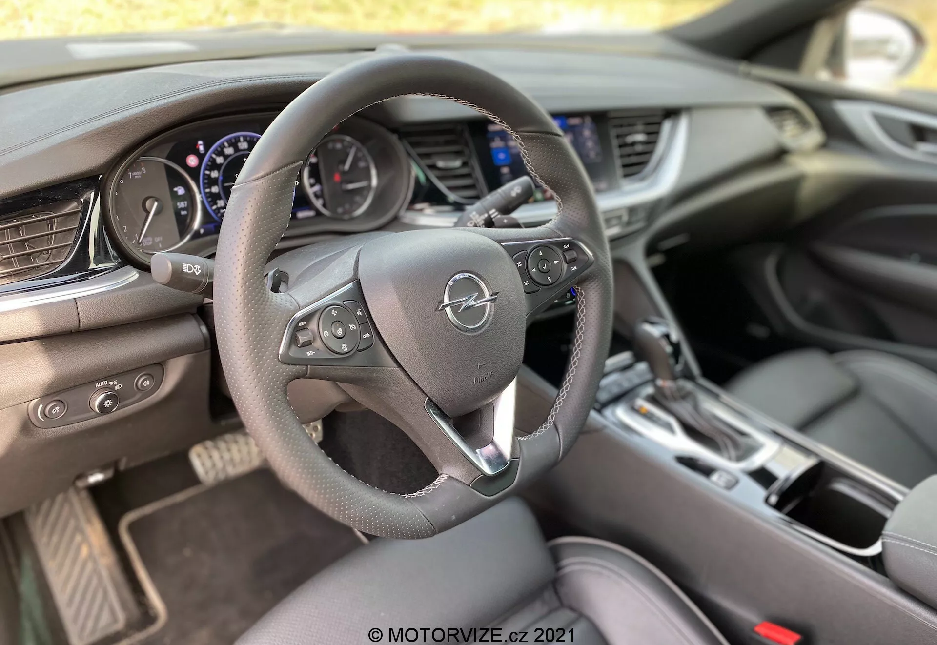 Interiörvy av en 2020 Opel Insignia GSI Grand Sport från förarens perspektiv, med fokus på en tre-ekrad läderklädd ratt med multifunktionskontroller, instrumentpanel med analoga mätare och en central digital display, en stor infotainmentskärm på mittkonsolen, horisontella luftventiler, klimatkontrollgränssnitt med fysiska knappar, växelspak för en automatisk växellåda, mugghållare och körlägesväljare på transmissionstunneln, allt belyst med naturligt dagsljus.