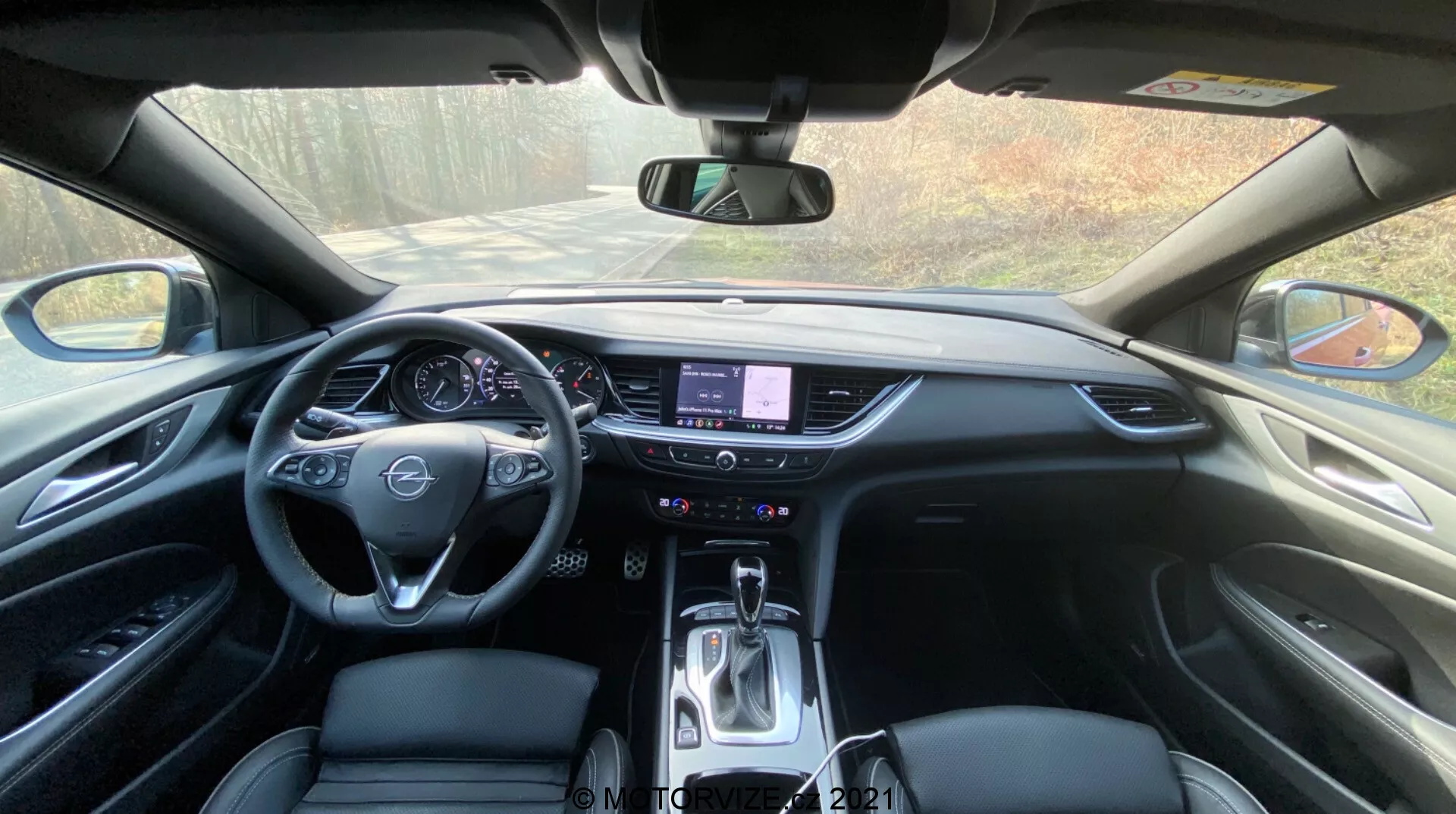 Interiör vy av en Opel Insignia GSi Grand Sport 2020 från förarens perspektiv, som visar instrumentbrädan med en centralt placerad ratt med inbyggda kontroller, en delvis synlig digital eller traditionell instrumentkluster, en framträdande mittkonsol med en pekskärmsinformationssystem, fysiska knappar och reglage för klimatkontroll under pekskärmen och en automatisk växelväljare. Den eleganta designen av instrumentbrädan integrerar mjuka plastmaterial, framhävda dekordelar och högkvalitativ stoppning, allt badat i naturligt dagsljus.